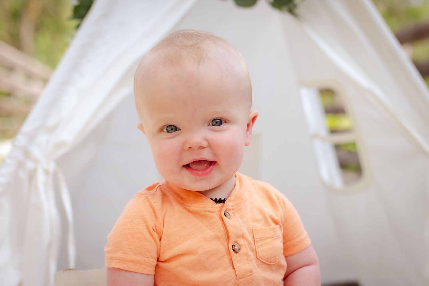 Baby in peach polo shirt smiles in front of white tent during outdoor photo session.