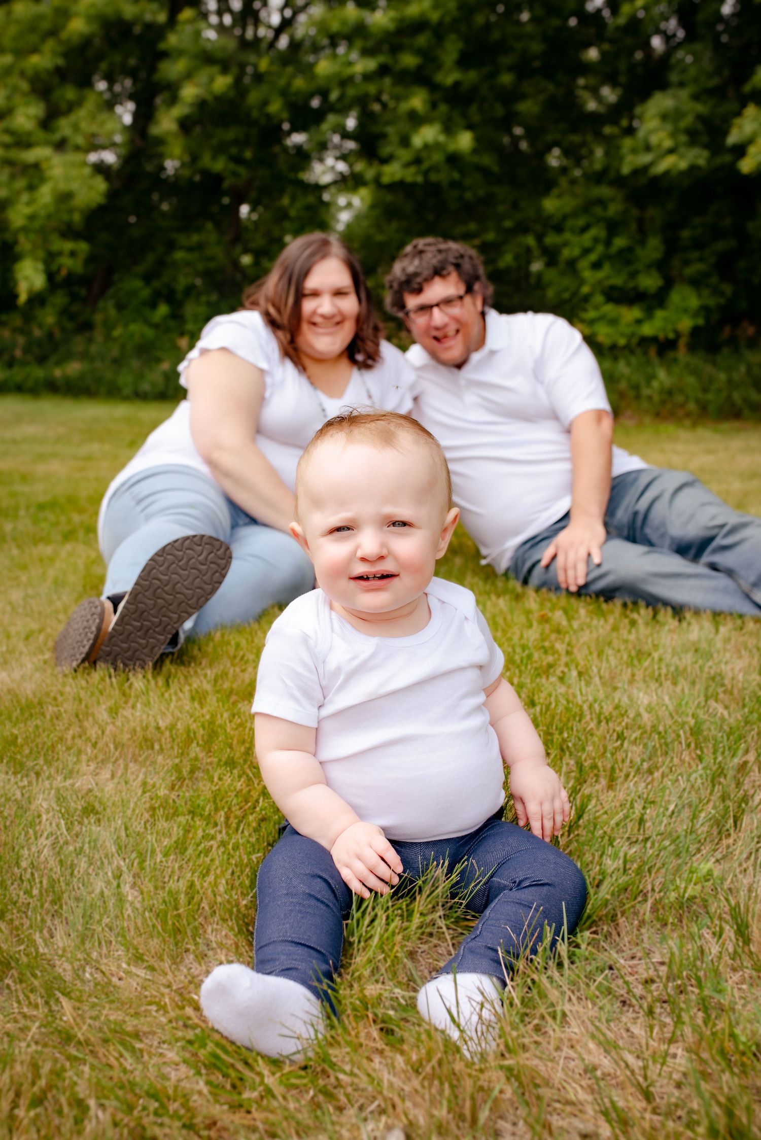 Toddler sits in grass while parents relax in background during outdoor photo session.