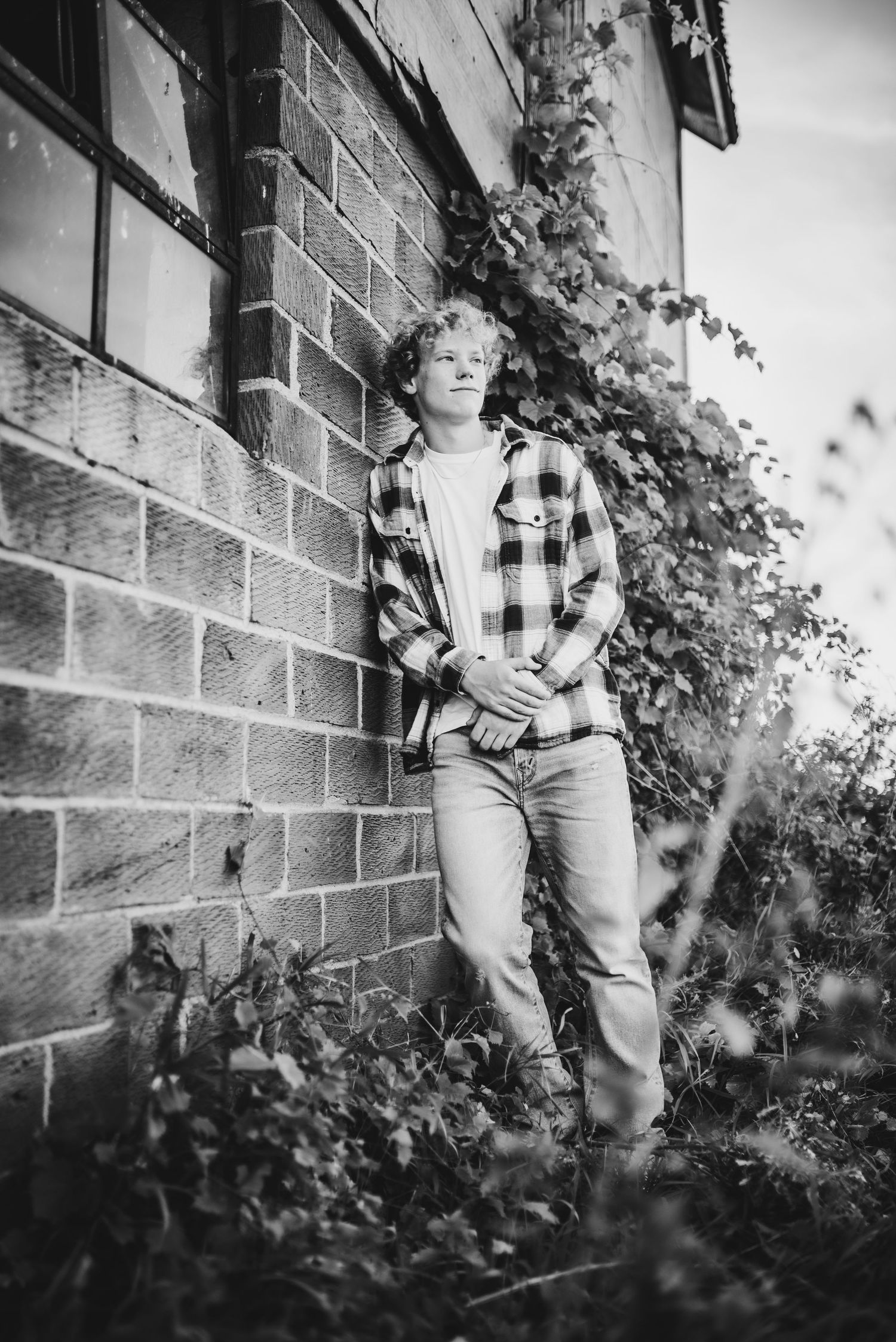 Black and white artistic shot against brick building with ivy, wearing plaid shirt and jeans.