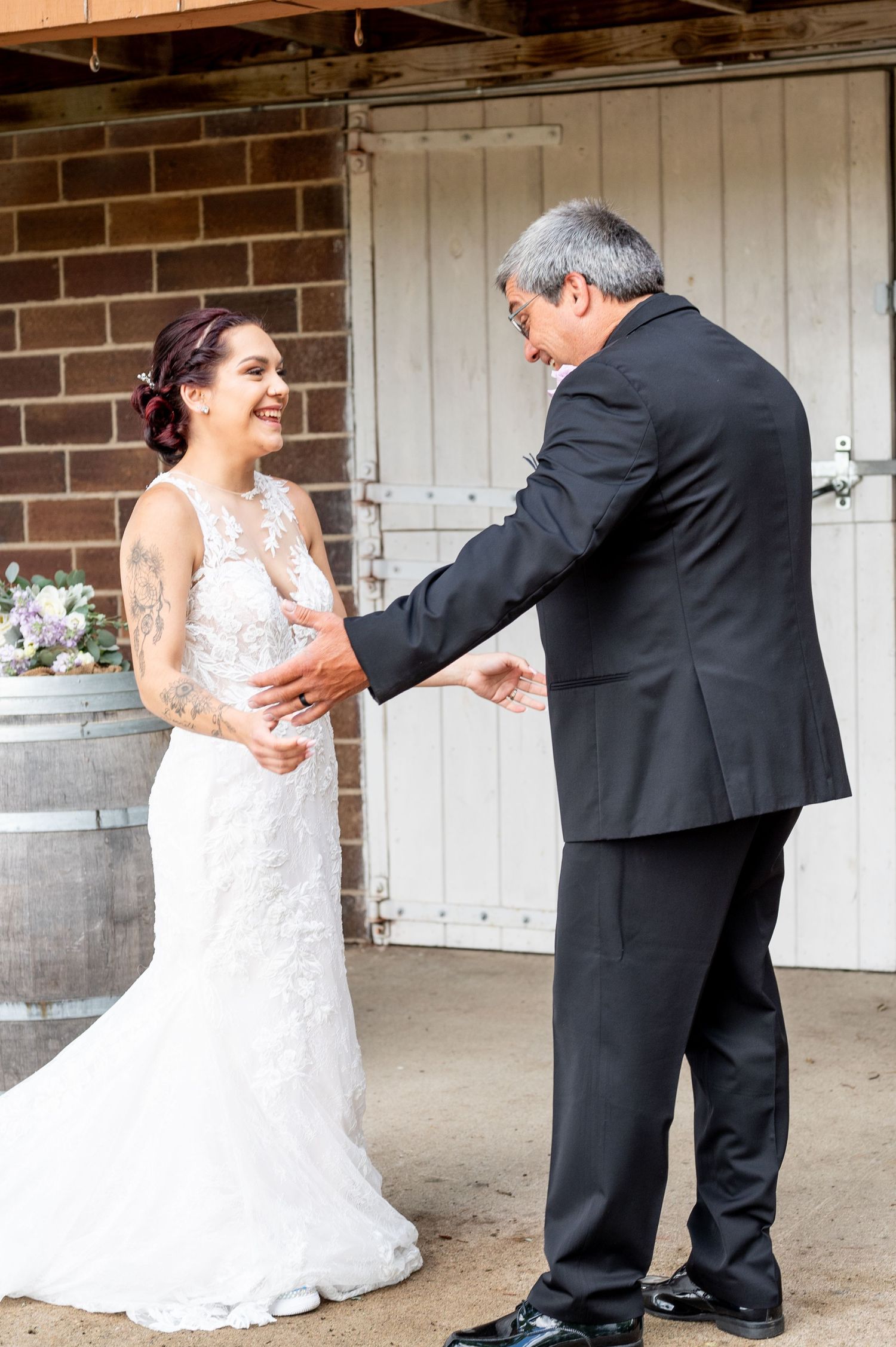 A joyful wedding moment between a bride and man in formal attire on a brick porch with rustic barrels.
