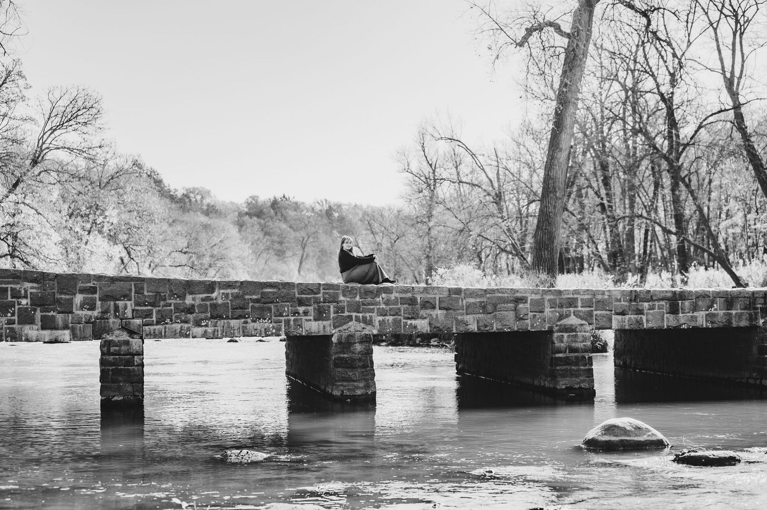 Black and white panoramic photograph showing a stone bridge spanning a river in winter with bare trees along the banks.