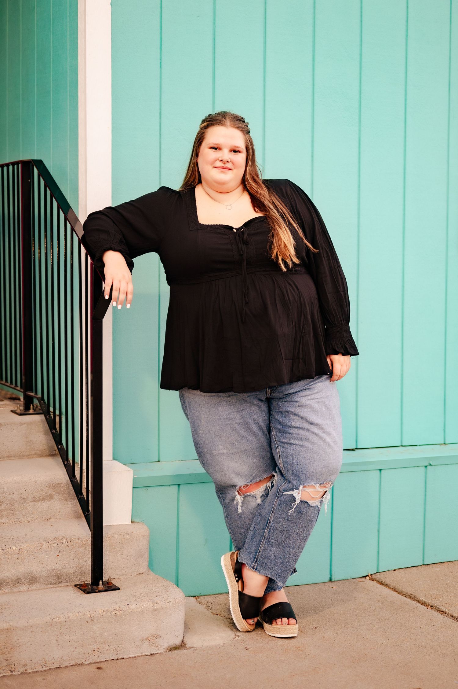 Person in black top and ripped jeans posing against turquoise wall with railing.