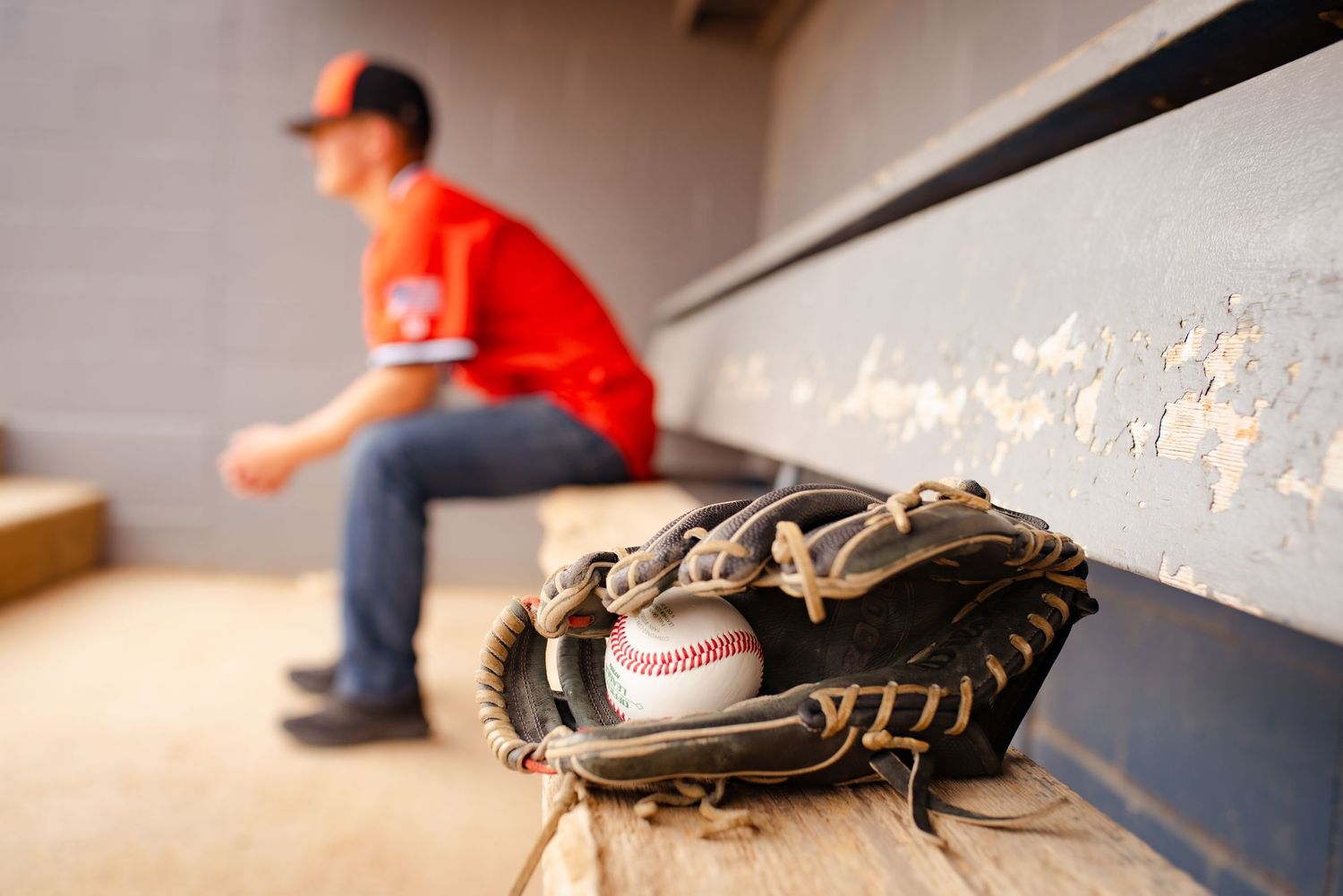 Baseball glove and ball rest on bench in dugout with player silhouetted in background.