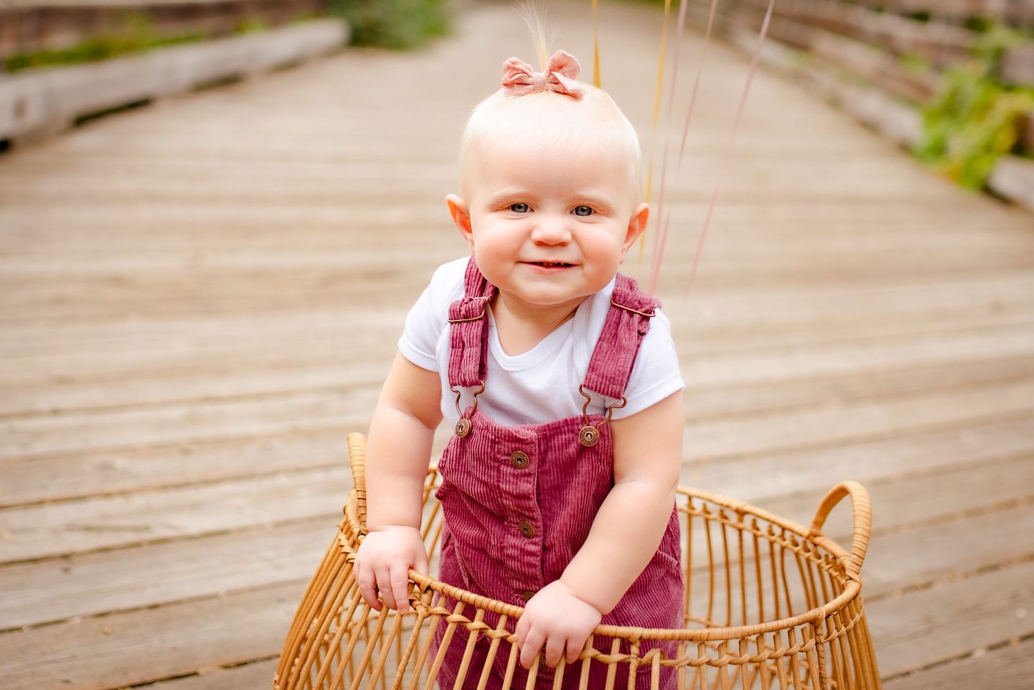 Baby wearing red overalls smiles while standing in wicker basket on wooden bridge.