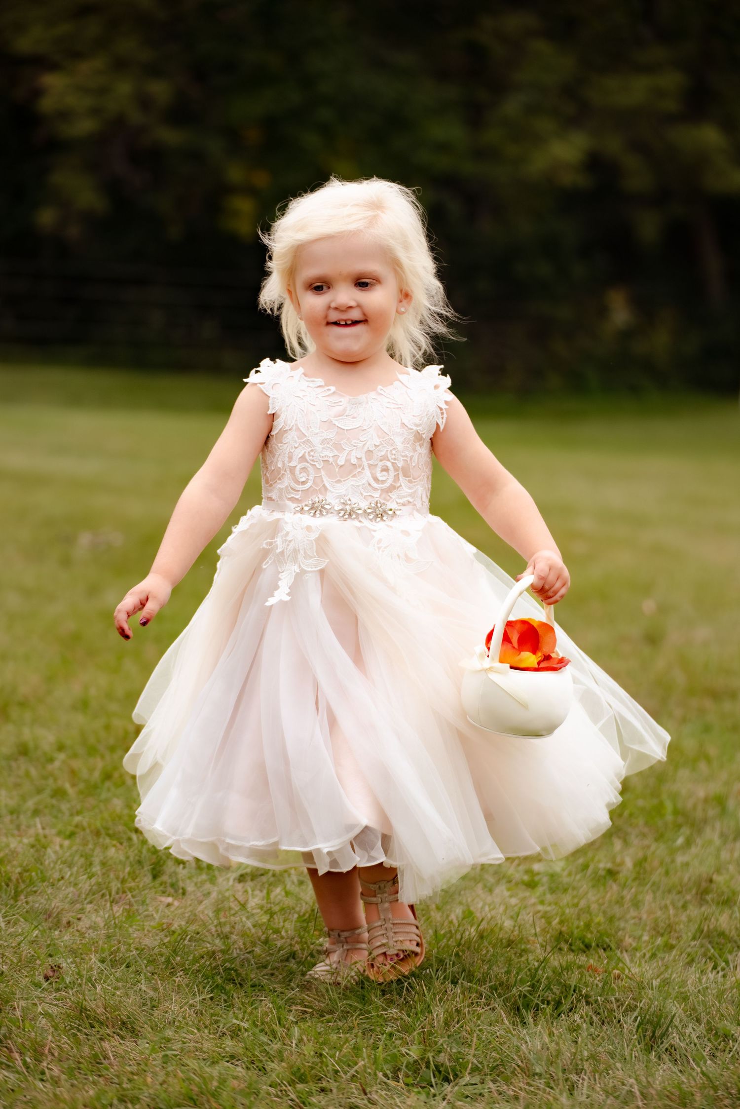 A young flower girl in a blush tulle dress skips through a grassy field carrying a white basket.
