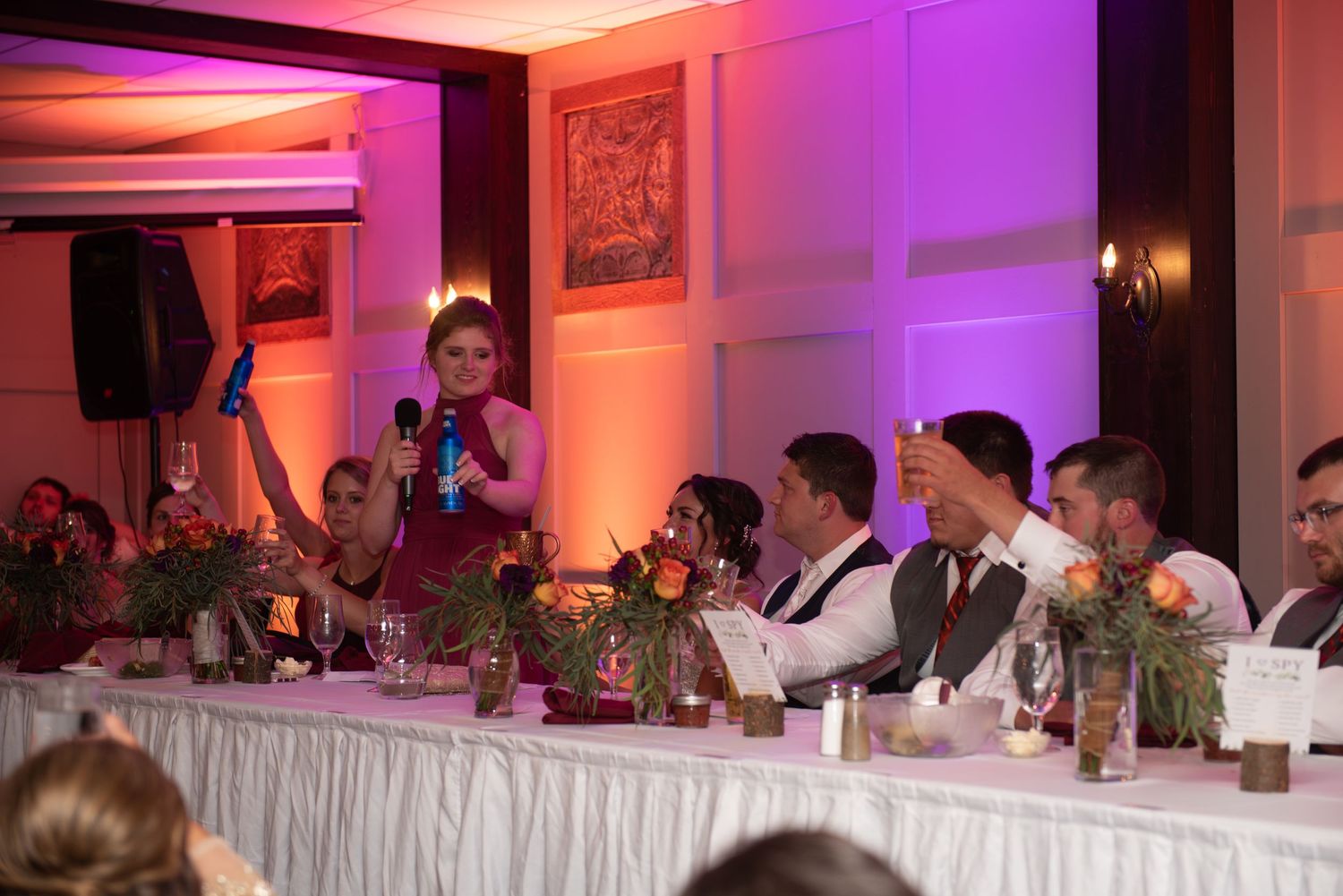 Wedding party members seated at a head table with colorful ambient lighting and flowers.
