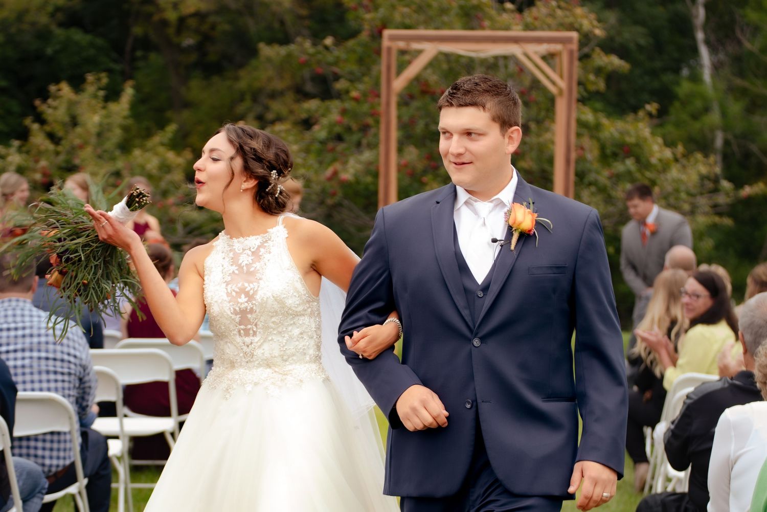 A couple celebrates during their outdoor wedding ceremony under a wooden arch with guests seated on white chairs.