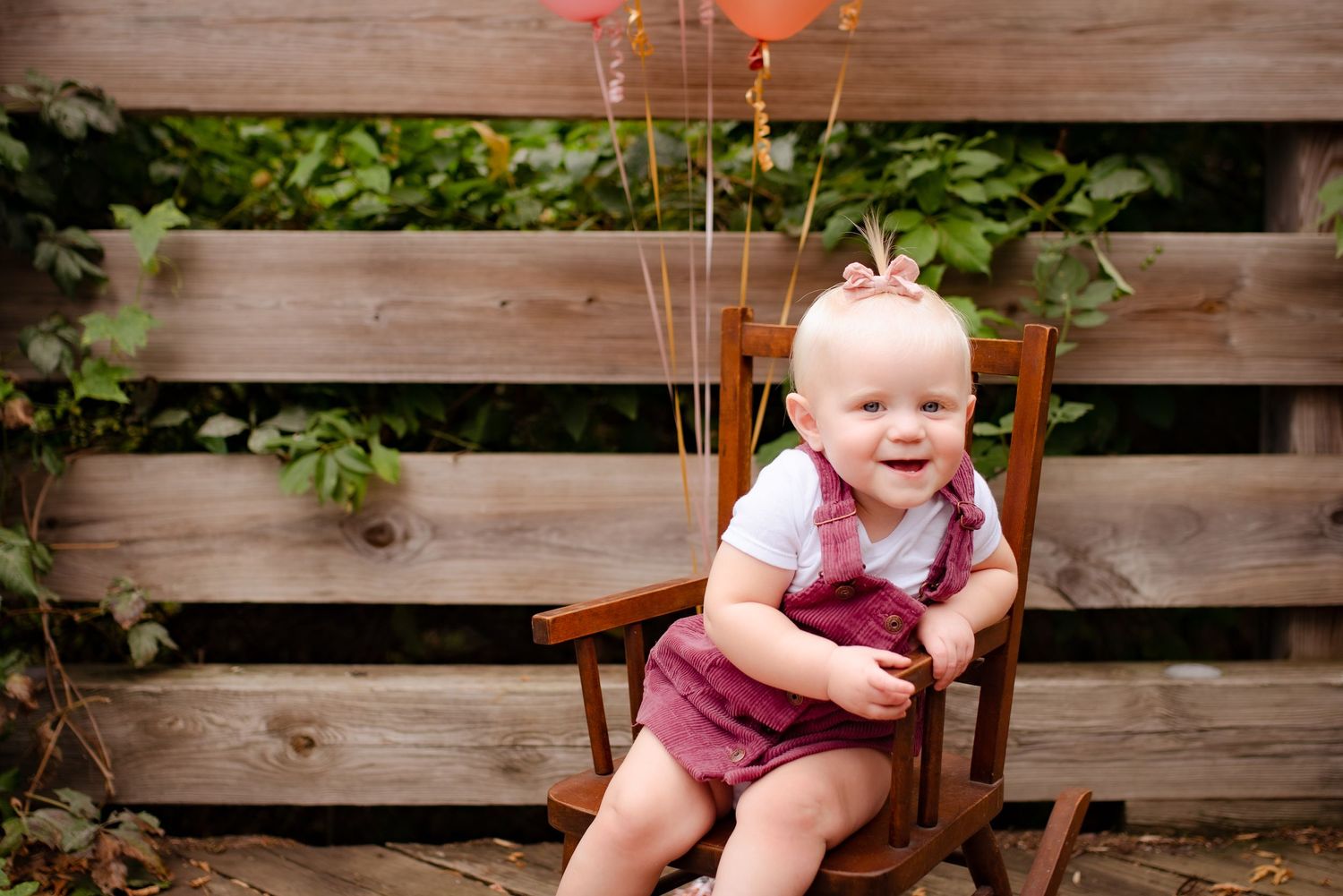 Baby sits in wooden rocking chair with balloons against rustic fence backdrop.