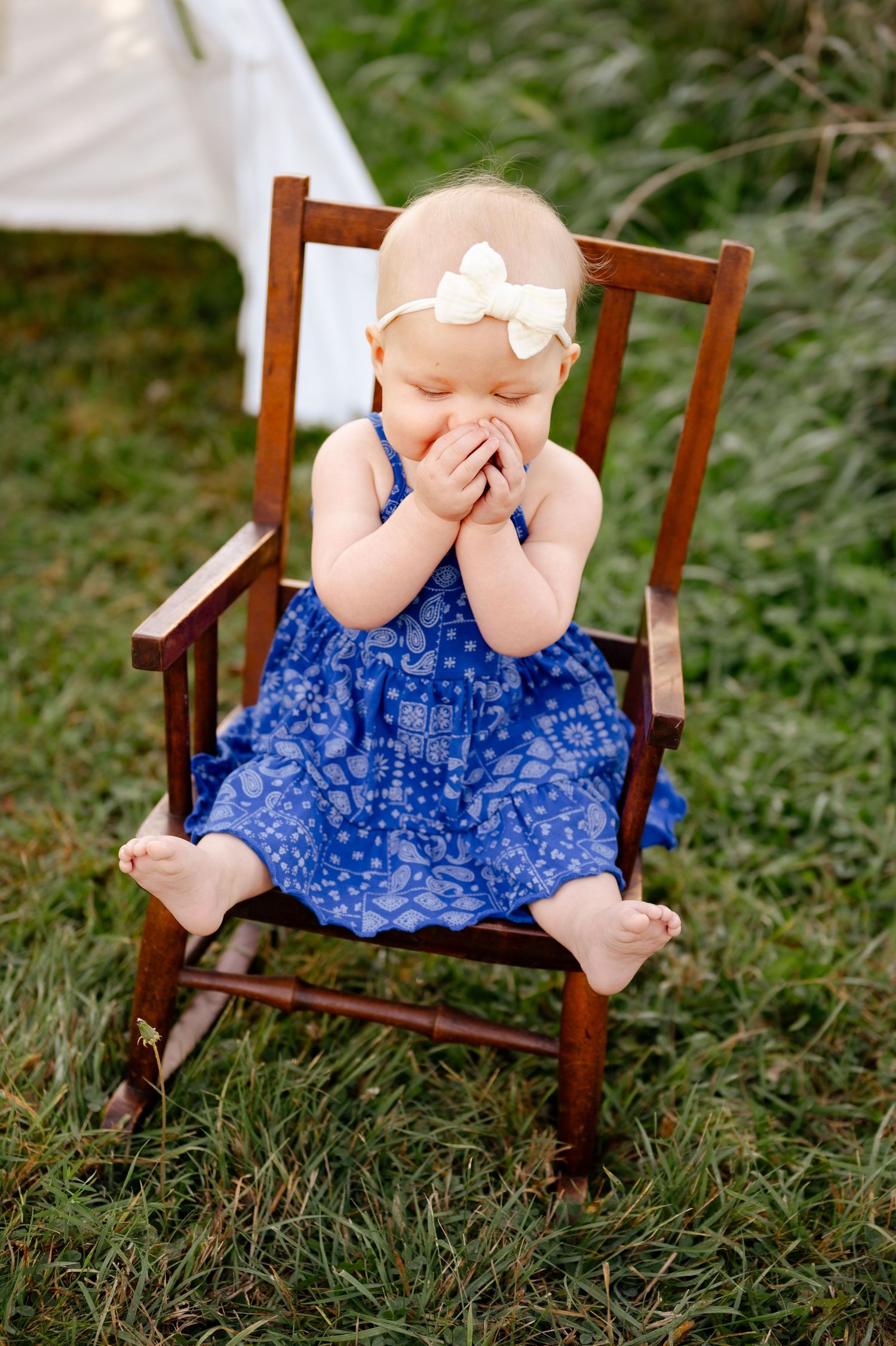 A toddler in a blue dress covers their face while sitting in a wooden rocking chair.