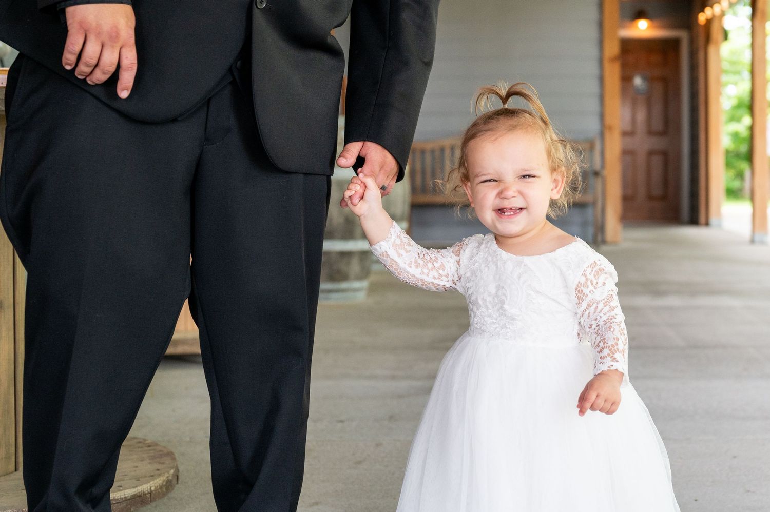 Adorable flower girl in white lace dress holding hands with someone in formal attire.