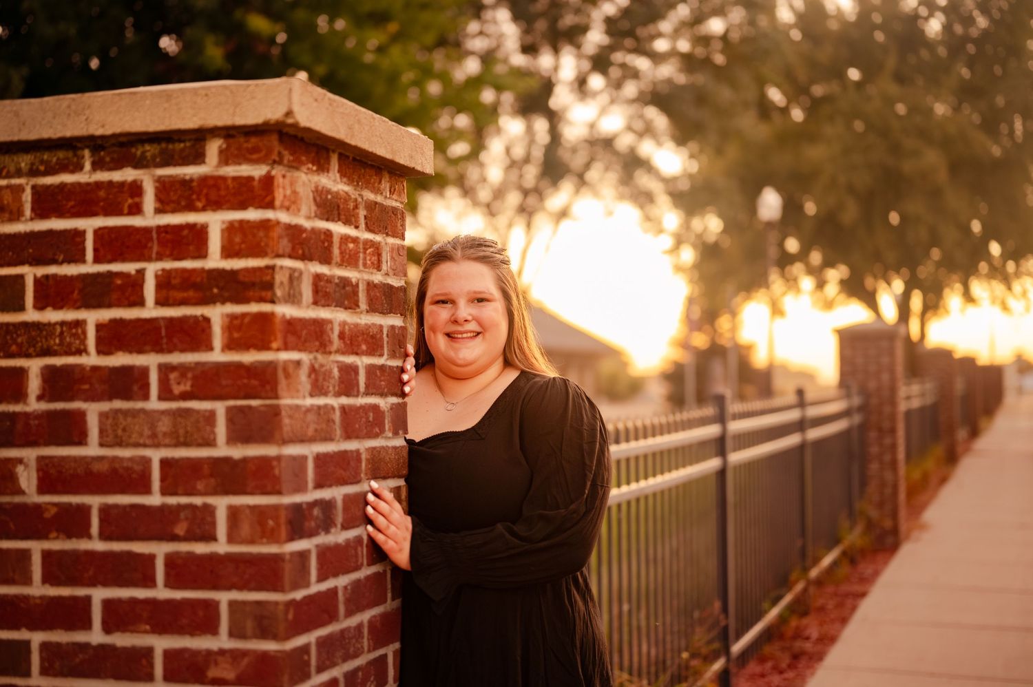 A person in a black dress poses next to a brick pillar at sunset with an iron fence in the background.