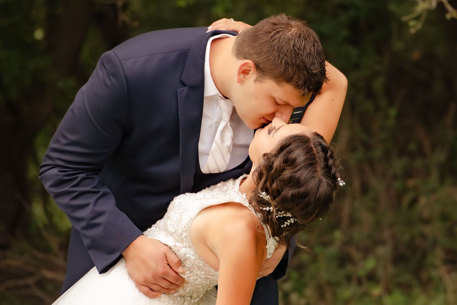 A romantic wedding kiss between newlyweds in formalwear against a natural outdoor backdrop.