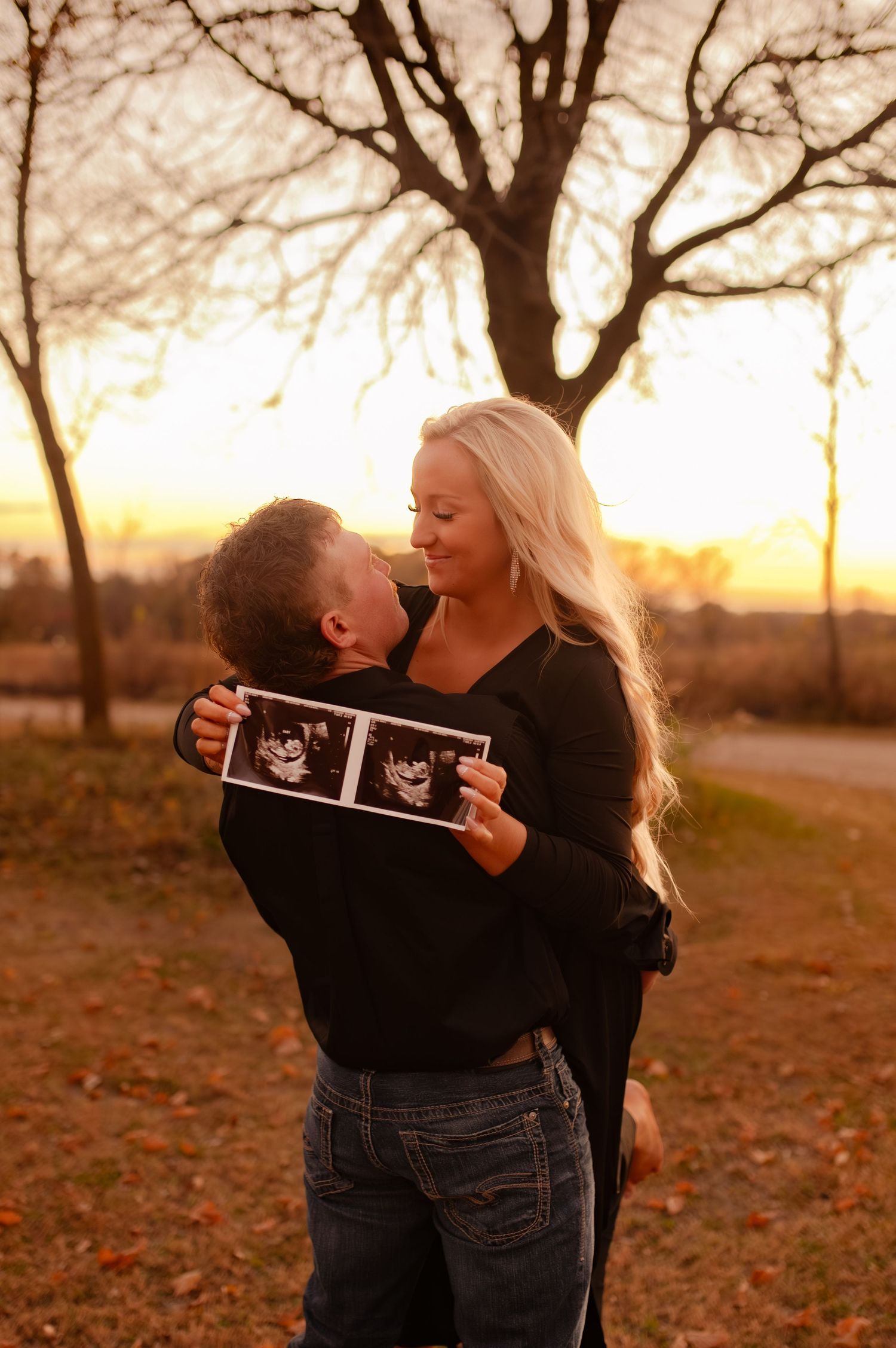 A couple shares an intimate moment in a park during sunset while holding ultrasound photos.