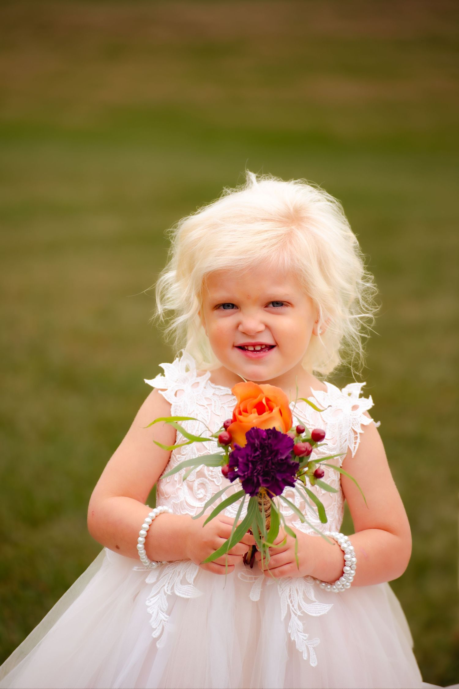A flower girl in white holds a small bouquet with orange and purple flowers against a grassy background.