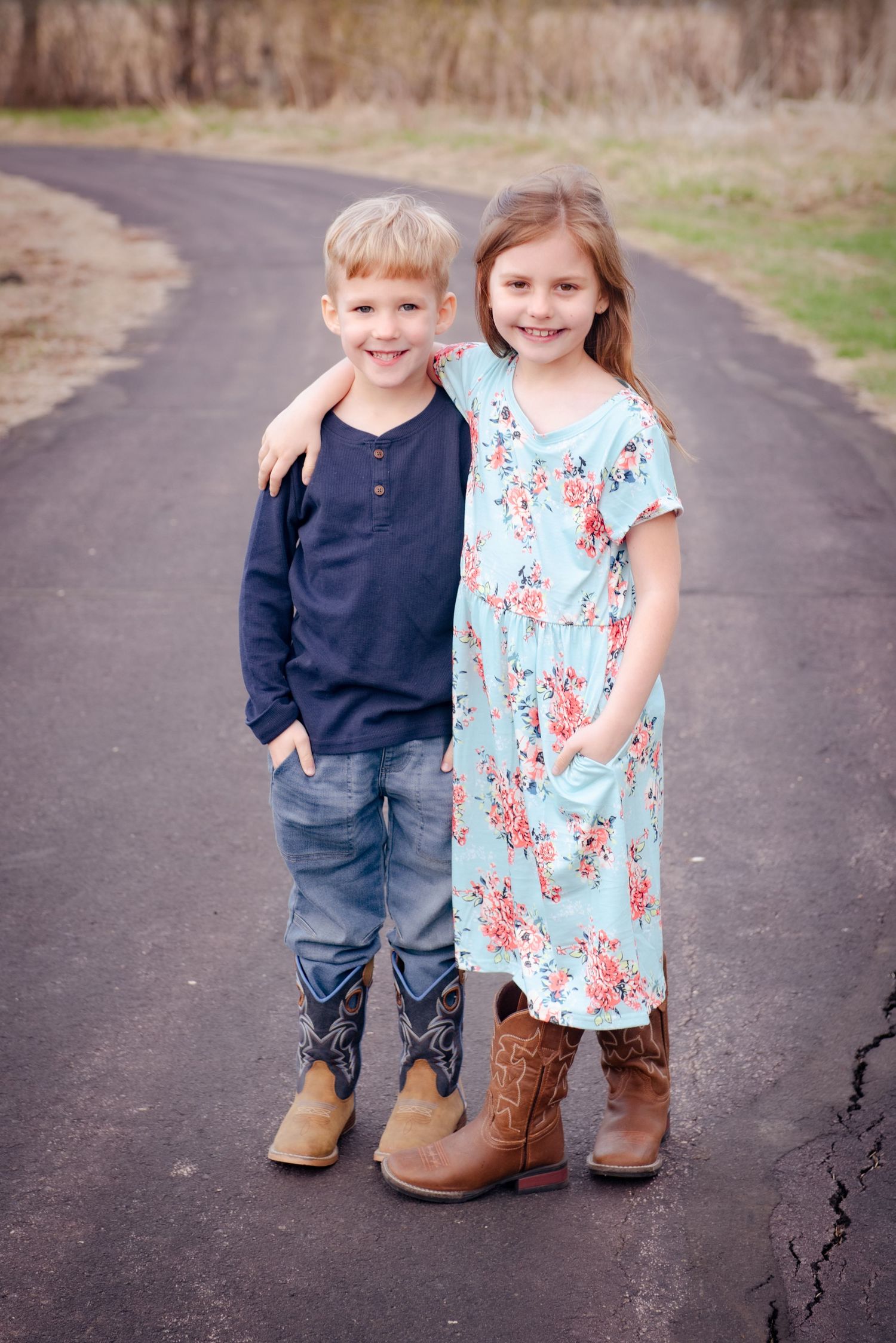 Two children standing together on a path wearing boots, one in a navy shirt and one in a floral dress.