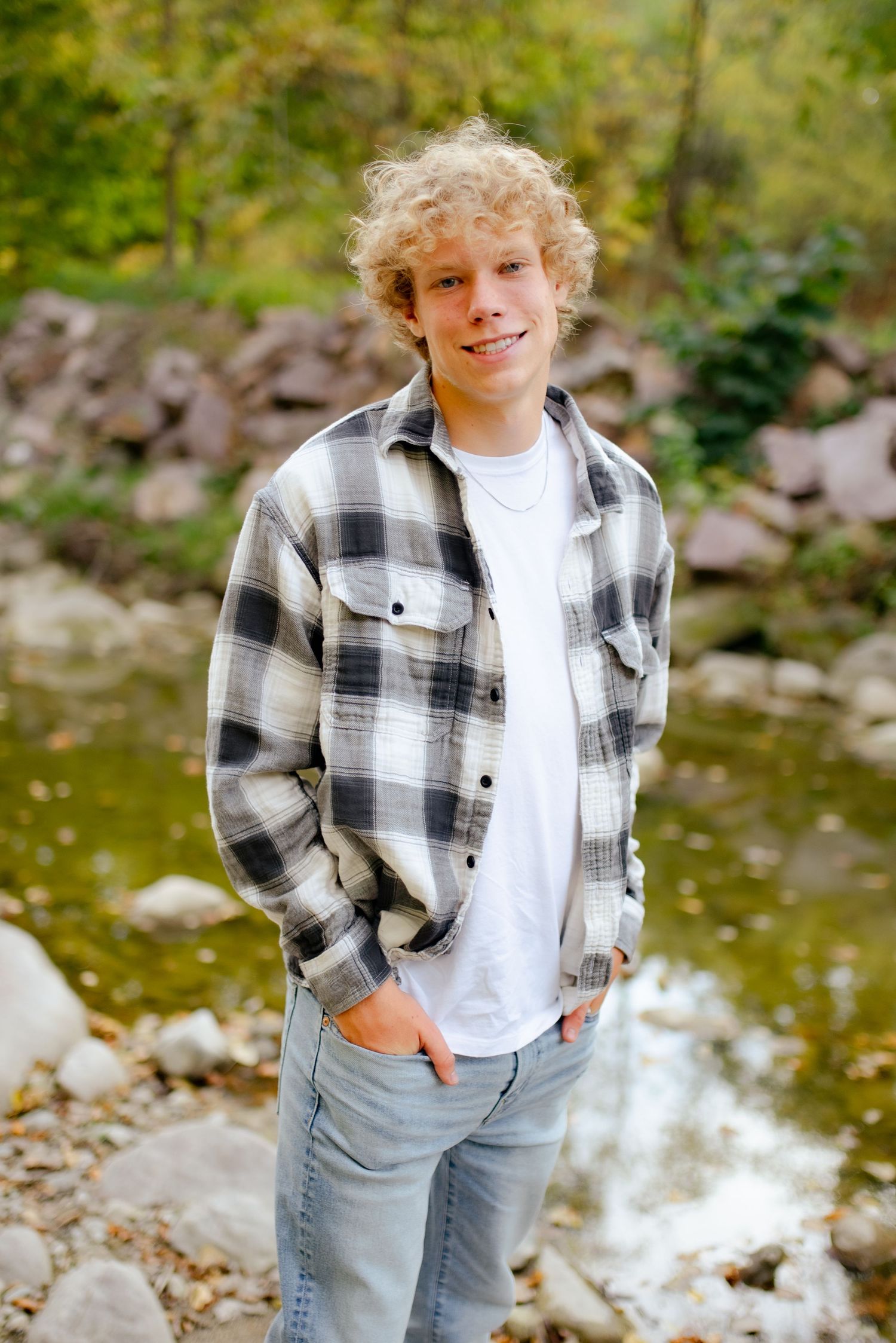 Someone in a plaid flannel shirt and jeans poses by a rocky stream in a natural setting.