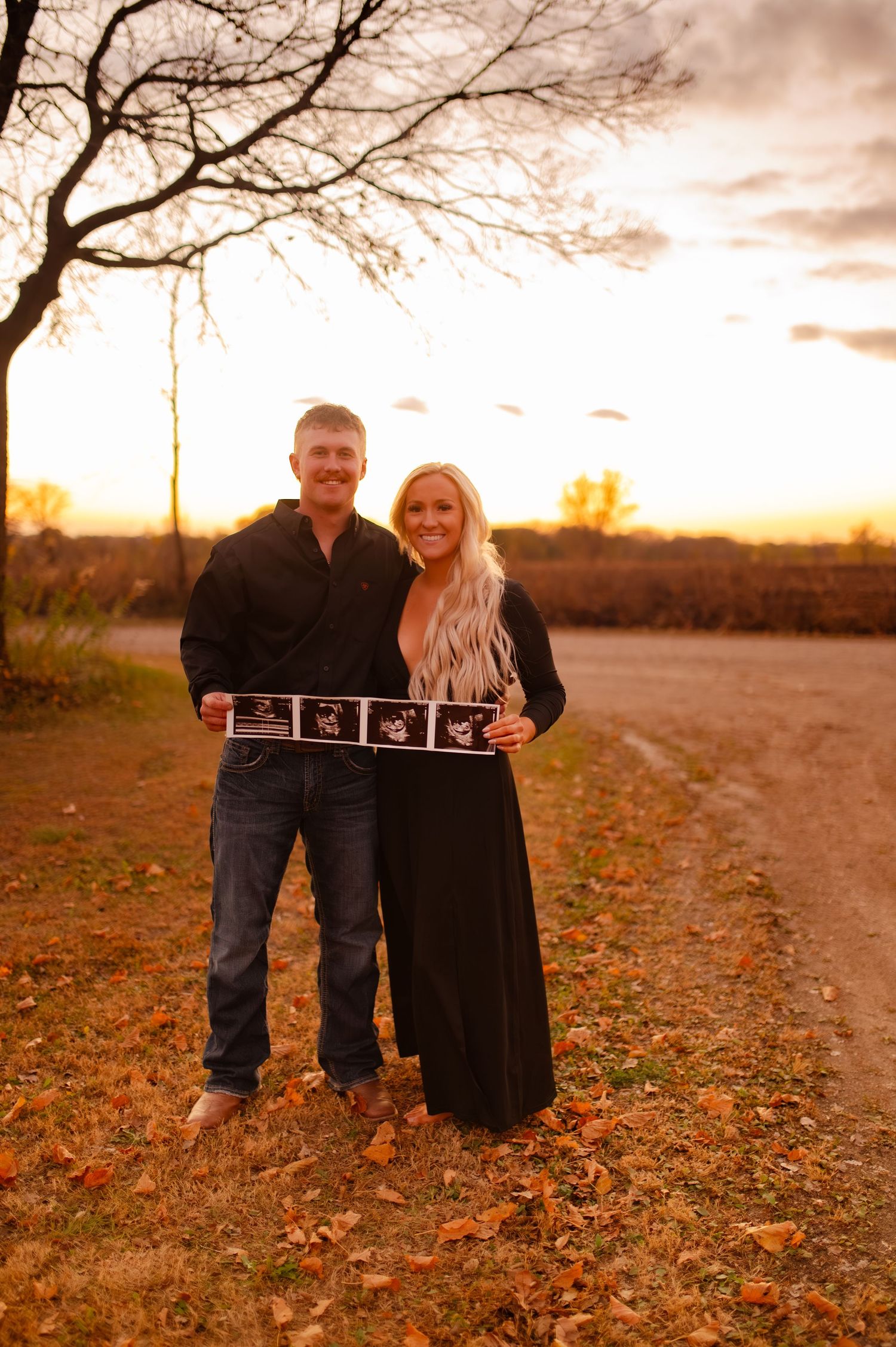 A couple poses together in a desert landscape at sunset while holding ultrasound photos in autumn.