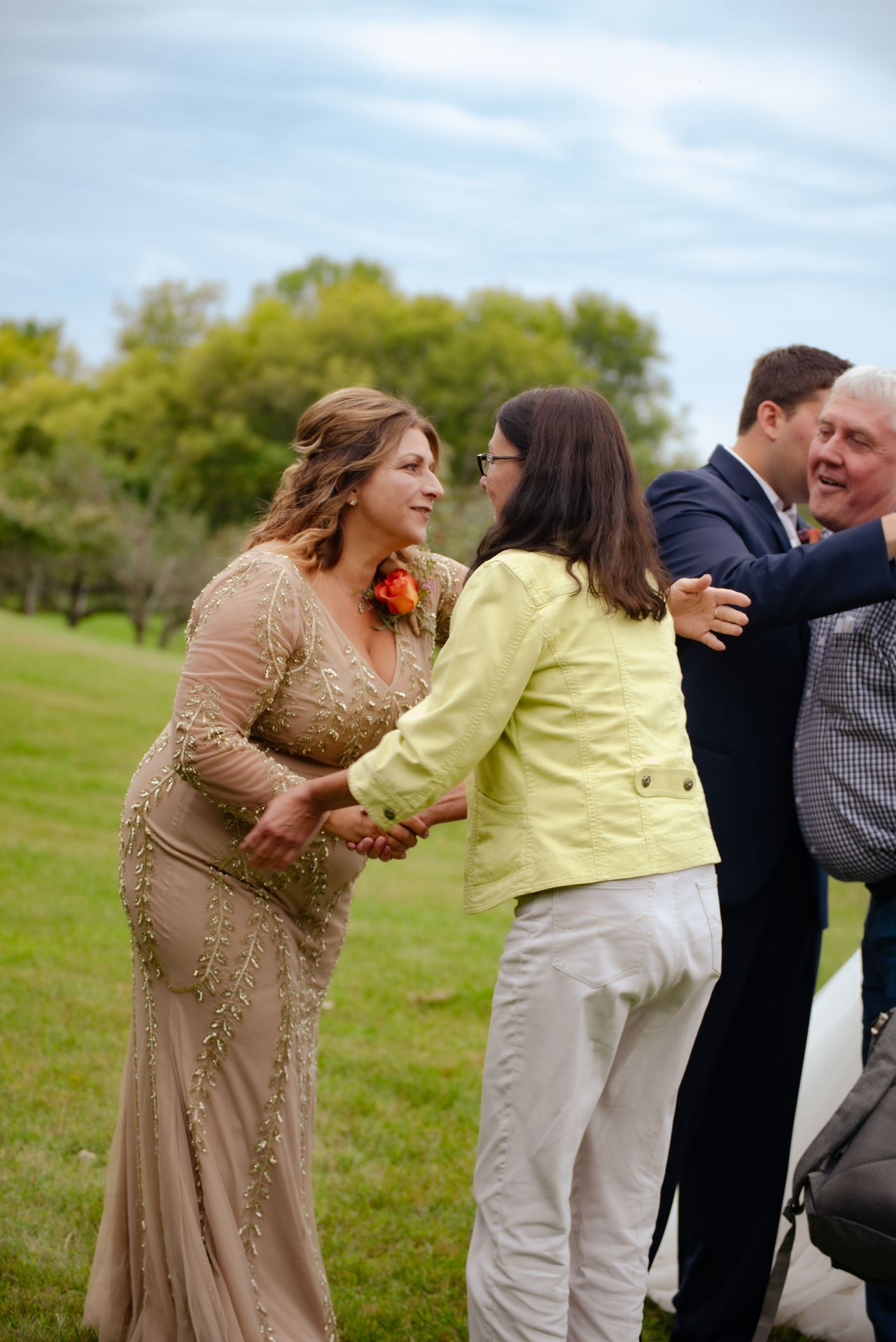 Guests greeting each other at an outdoor event with one wearing a beige dress and another in a yellow jacket.