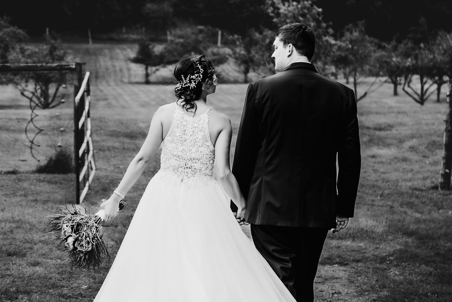 Black and white image of newlyweds walking away together through an orchard landscape.