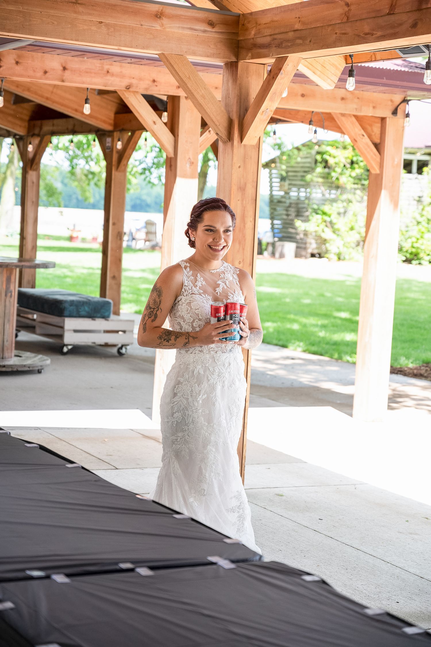 A bride in a lace wedding gown holding a drink under a wooden pavilion structure.