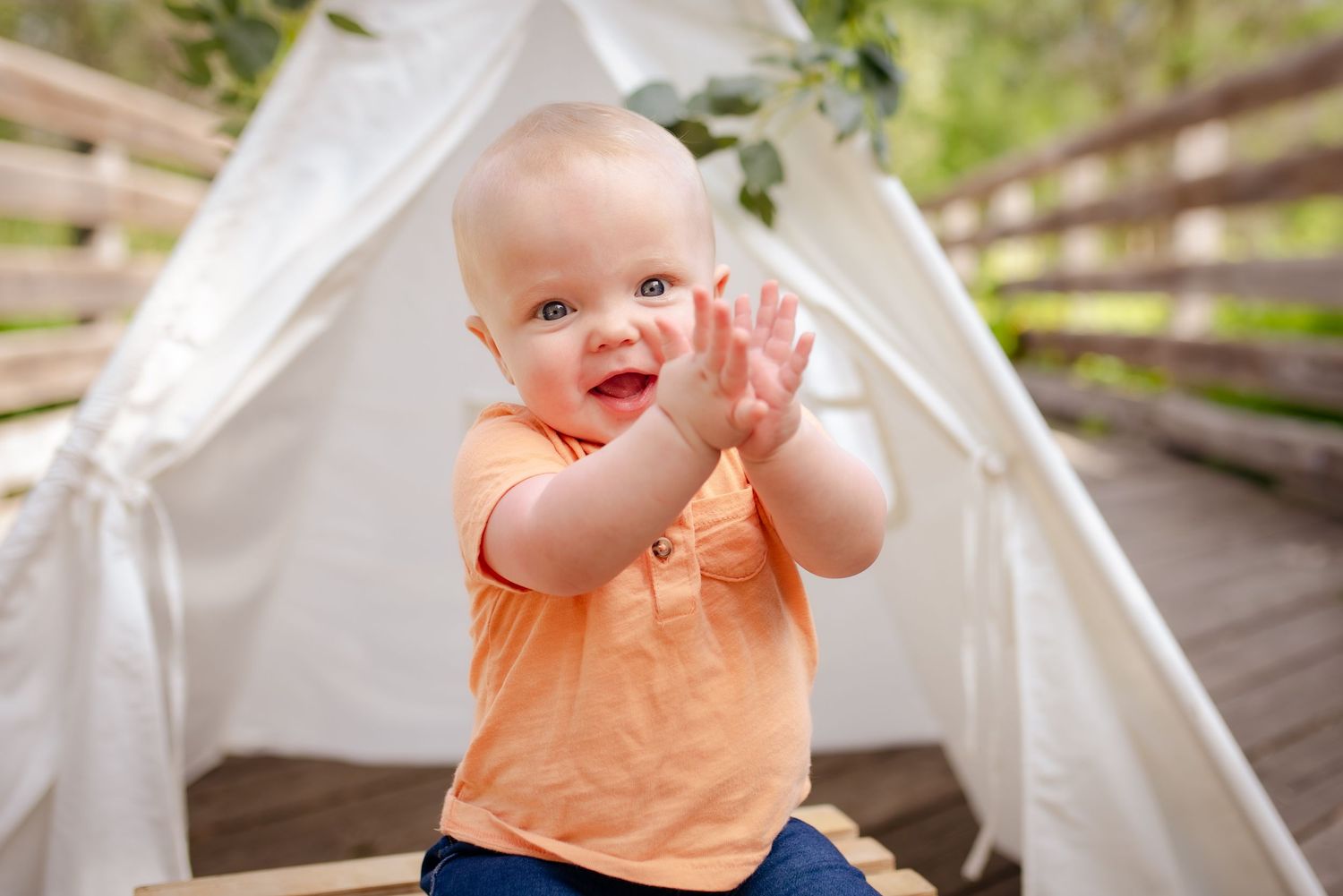 A joyful baby in orange shirt claps hands while sitting in a white tent.