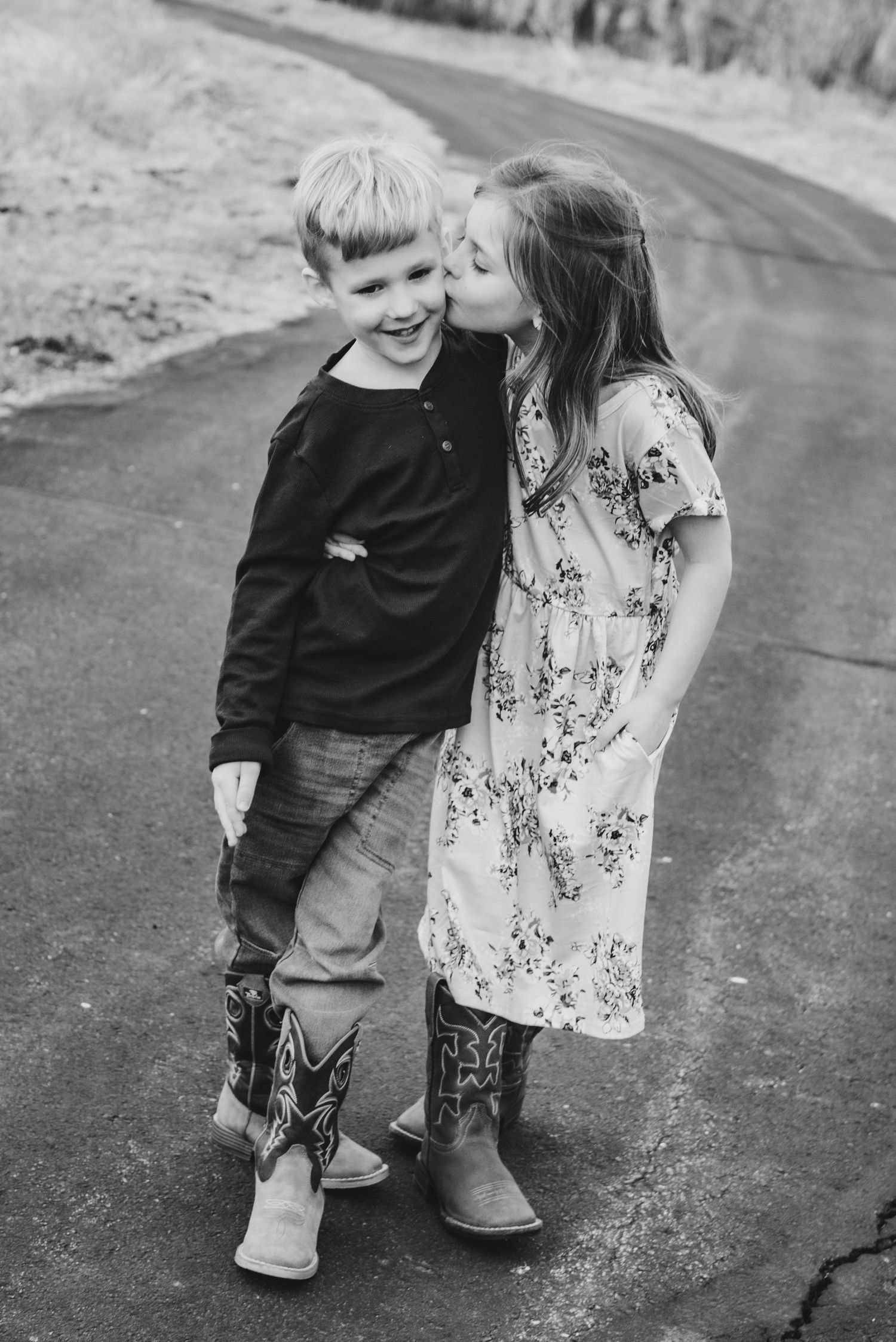 Black and white photo of two young children sharing a playful moment on a path.