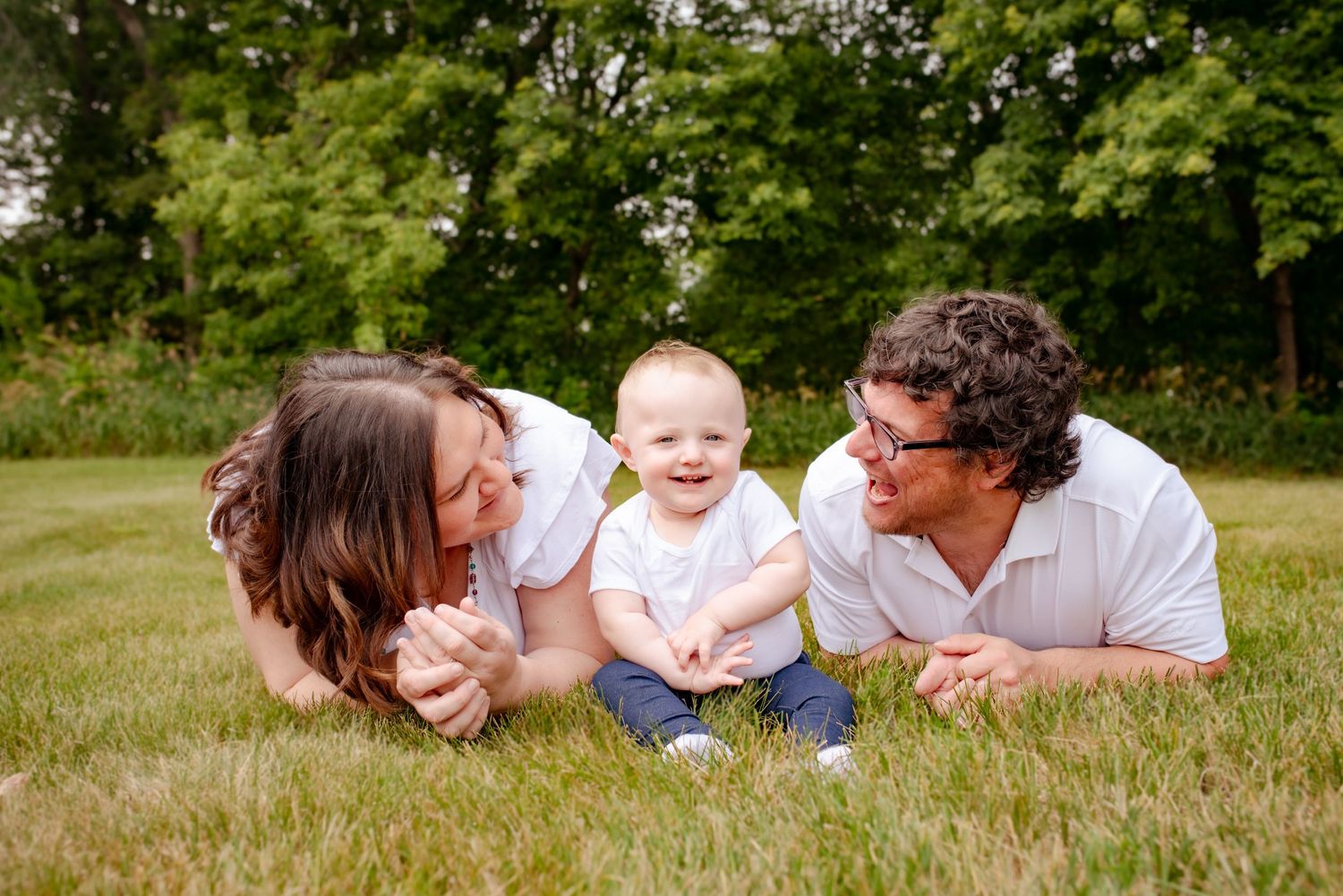 A family of three laughs together while relaxing on grass during an outdoor photo session.