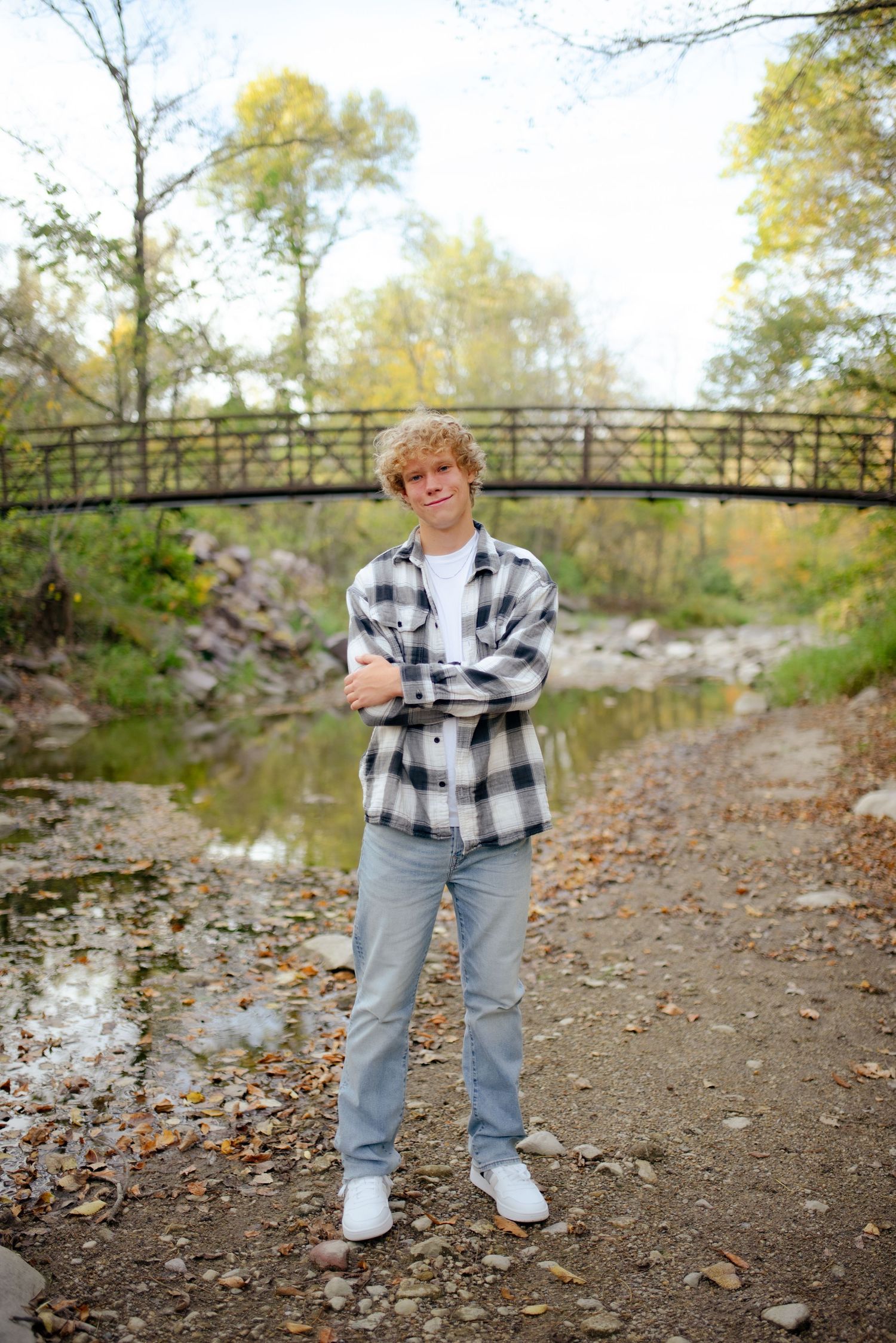 Someone in casual attire poses in front of a metal bridge over a stream during autumn.