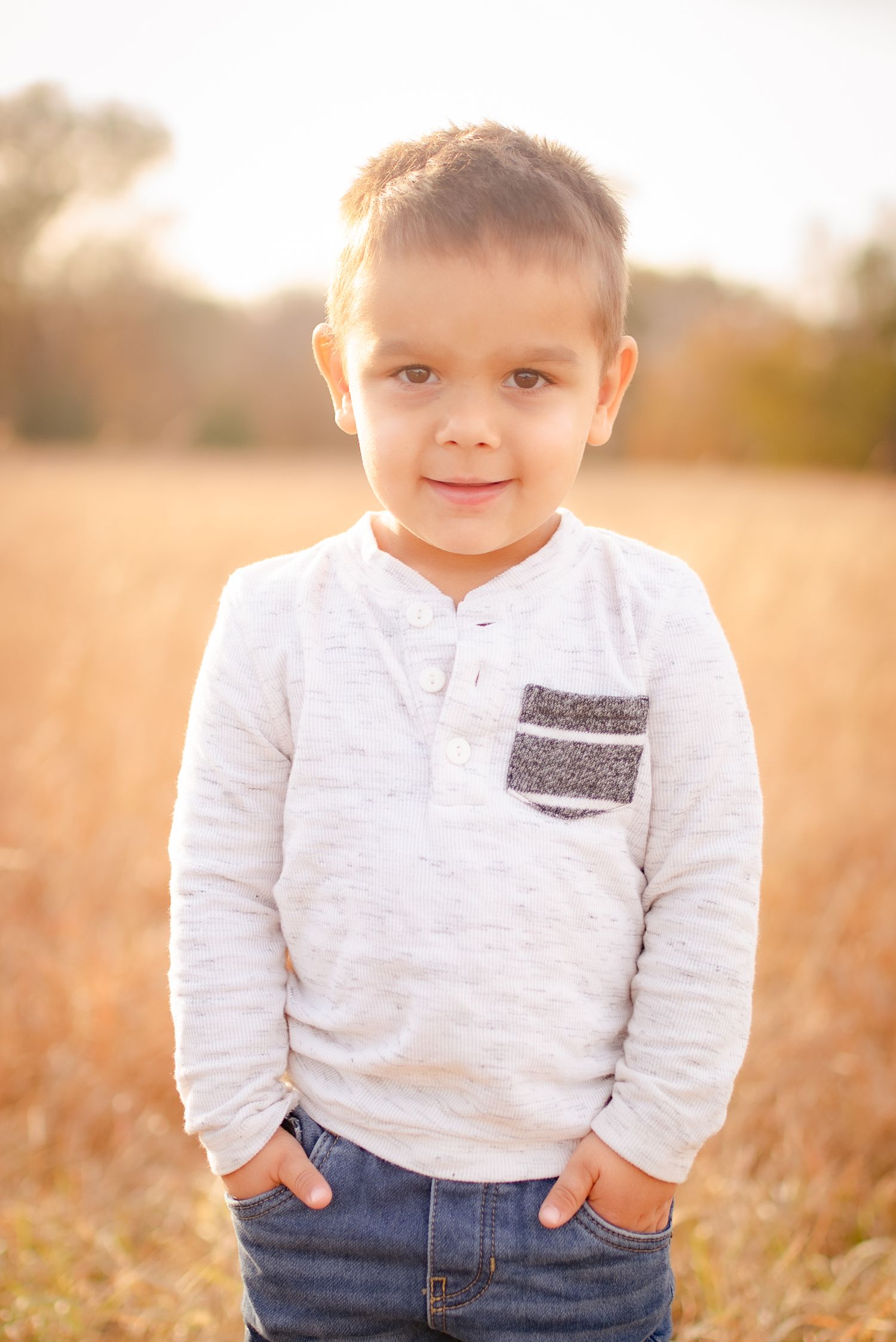 Child wearing a white henley shirt poses in a golden wheat field at sunset.