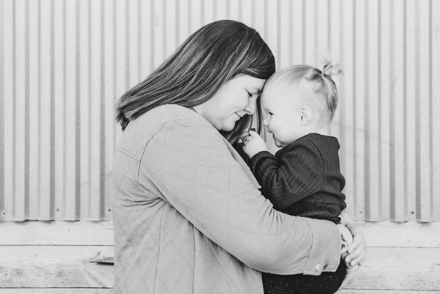 Intimate black and white portrait of mother and baby sharing a tender moment against metal wall.