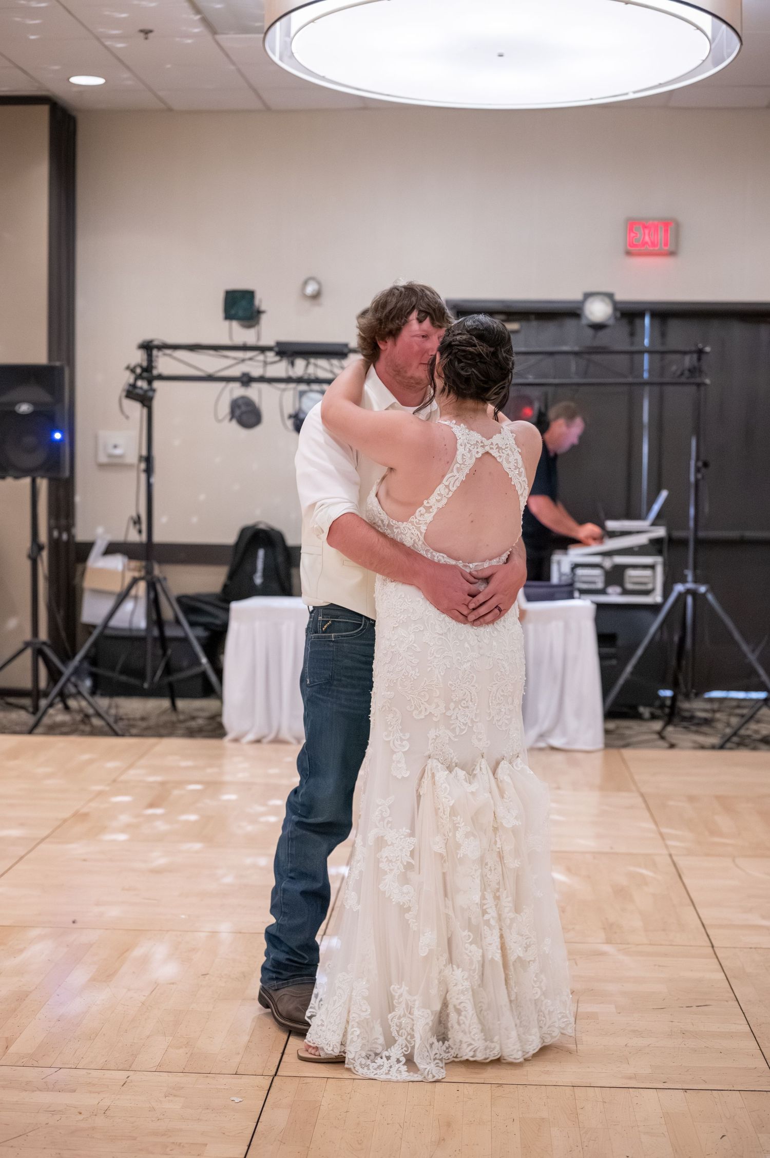 A couple shares their first dance at a wedding reception on a wooden dance floor with DJ equipment in the background.