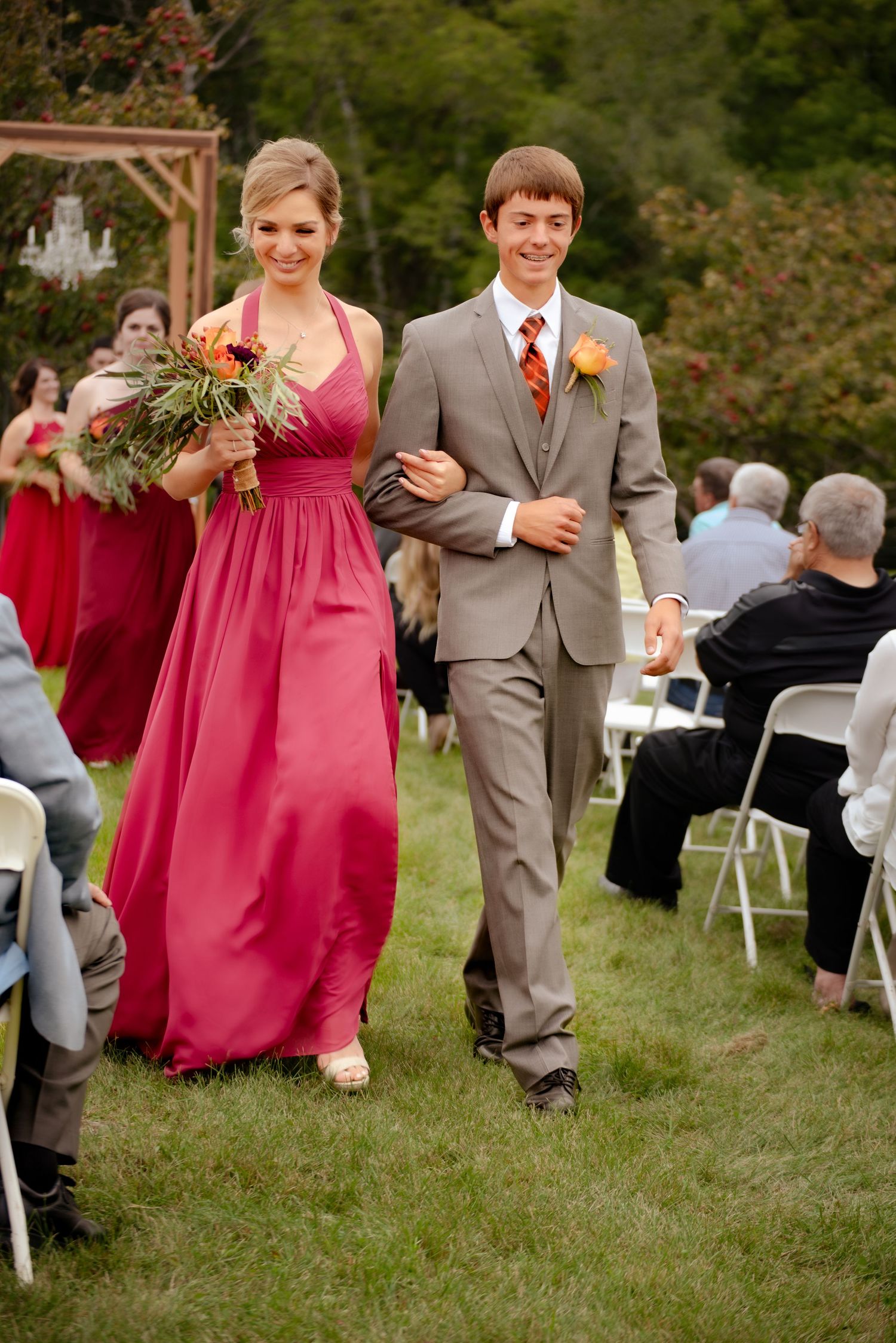 Wedding party members walk down an outdoor aisle with bridesmaids in red dresses and groomsmen in gray suits.