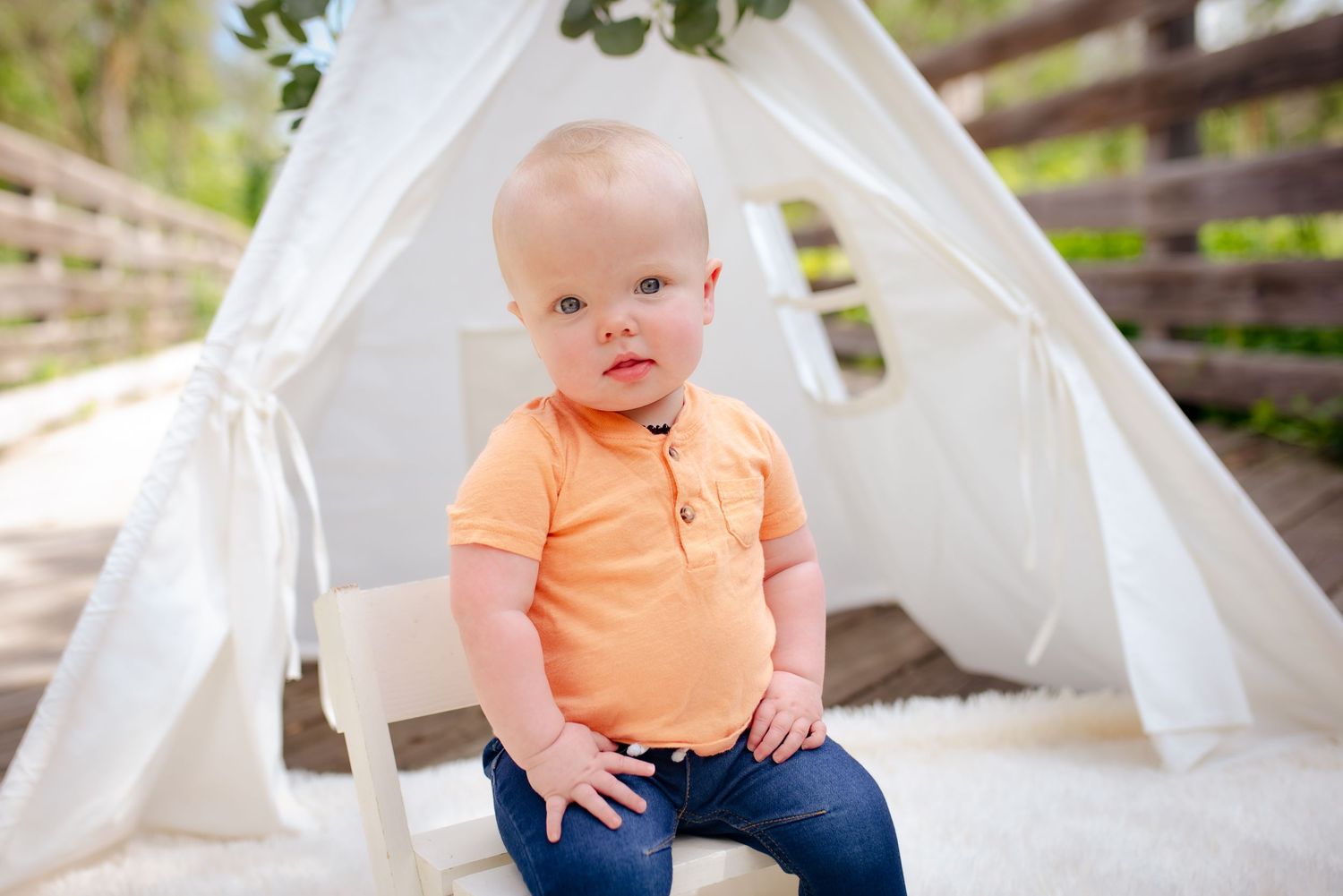 Baby wearing orange polo shirt poses in front of white teepee tent outdoors.