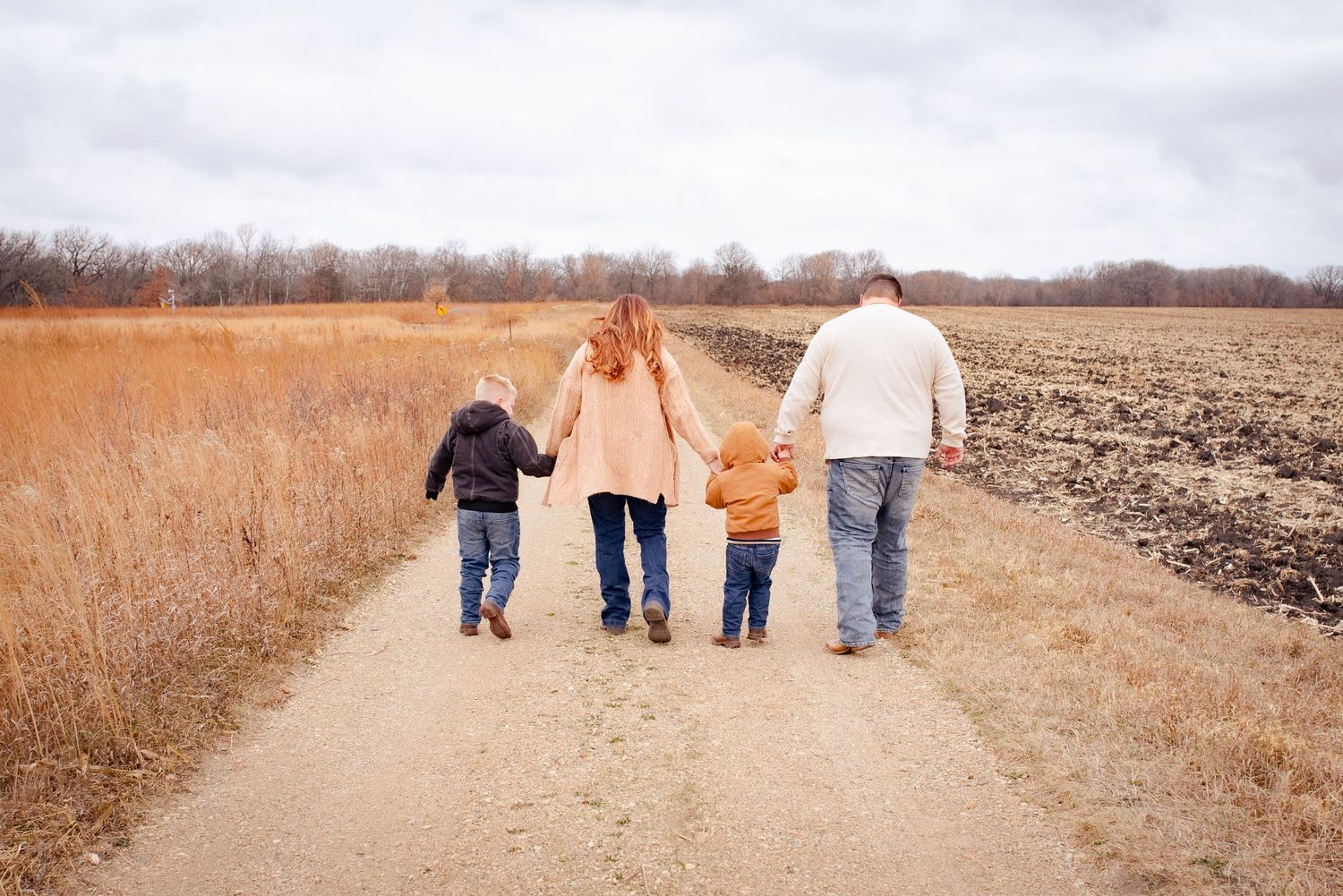 Family walking hand in hand down rural dirt path surrounded by autumn fields.