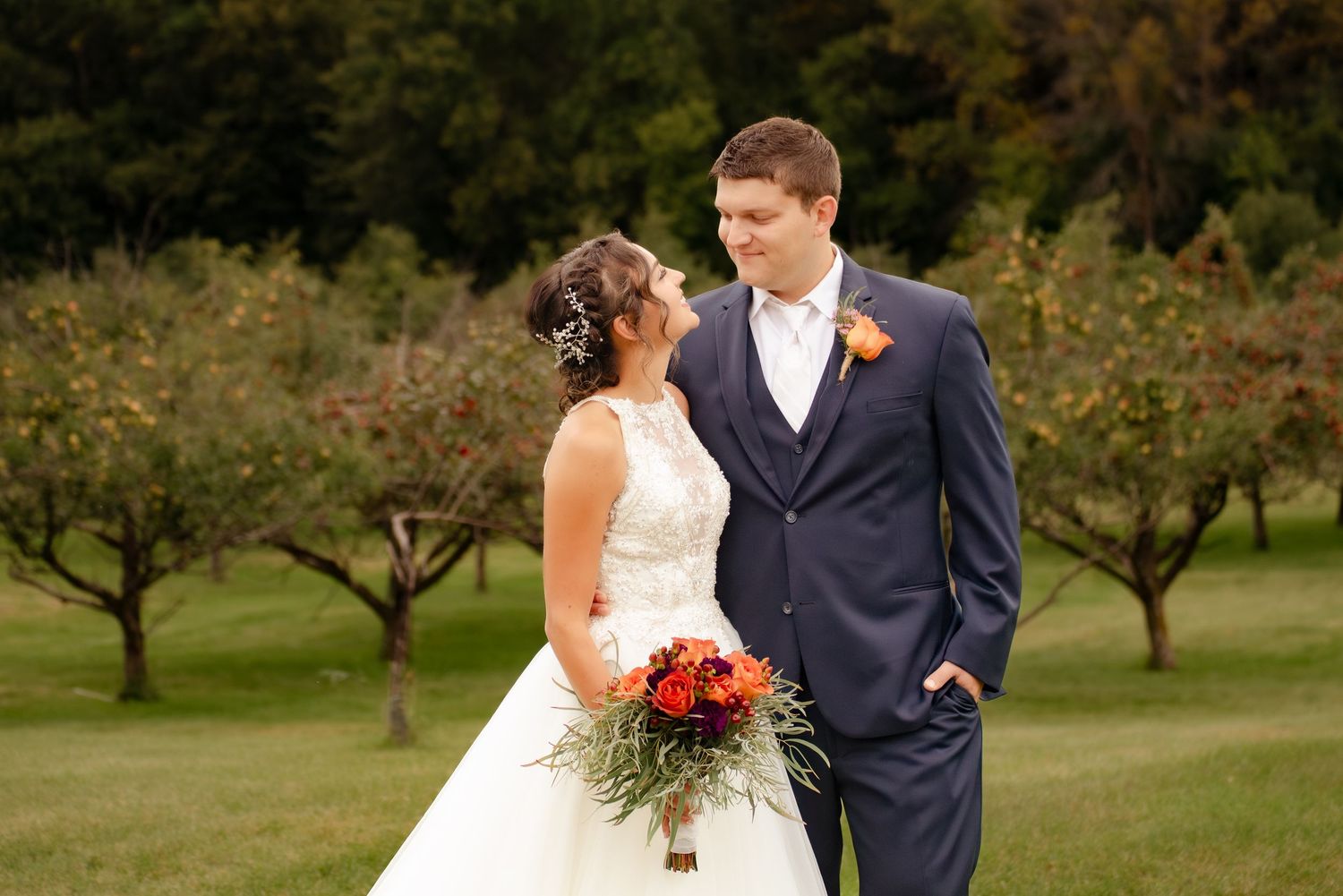 Newlyweds in formal attire pose together in an orchard with the bride holding a vibrant autumn bouquet.