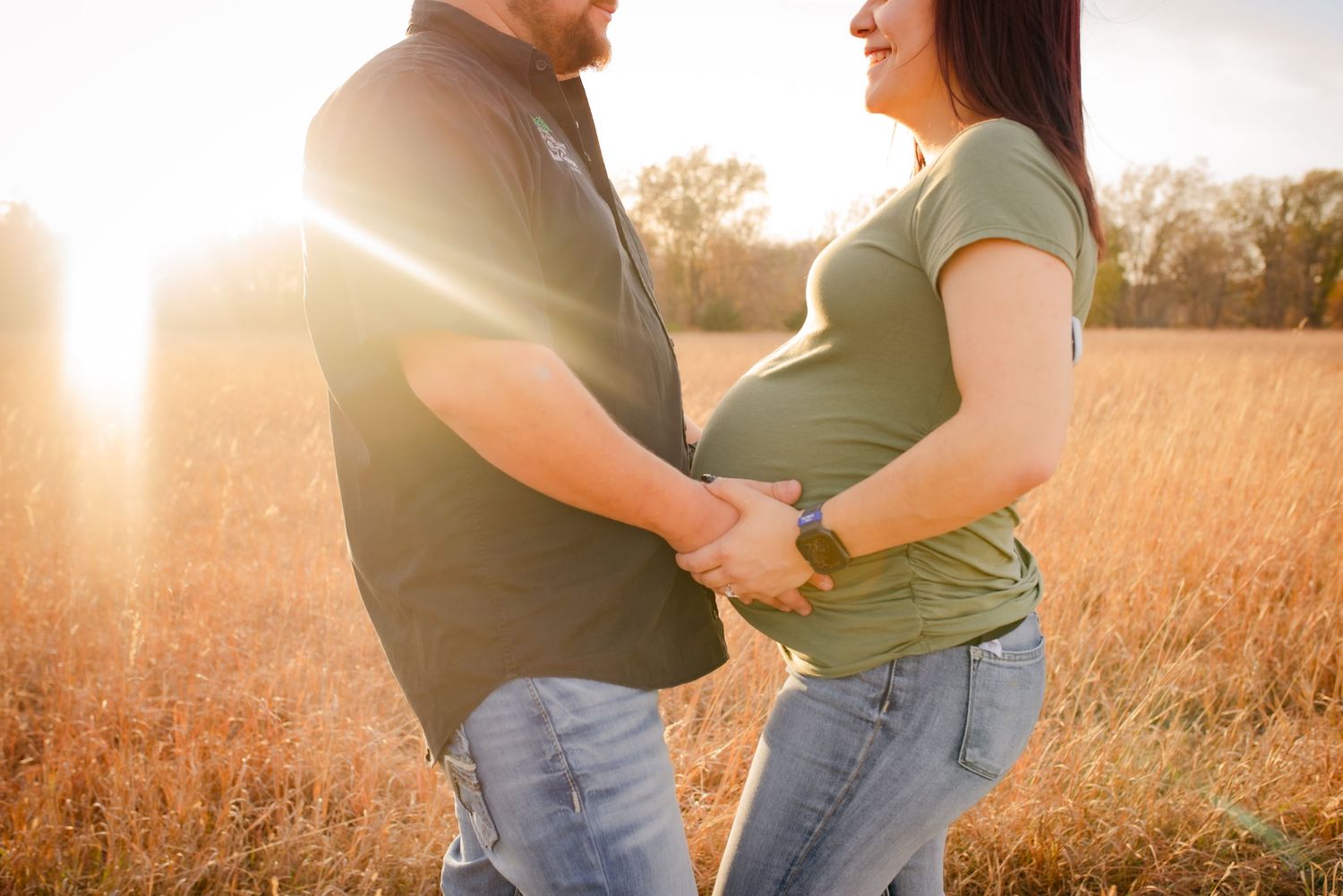 Side-lit maternity portrait of couple holding hands in golden field.