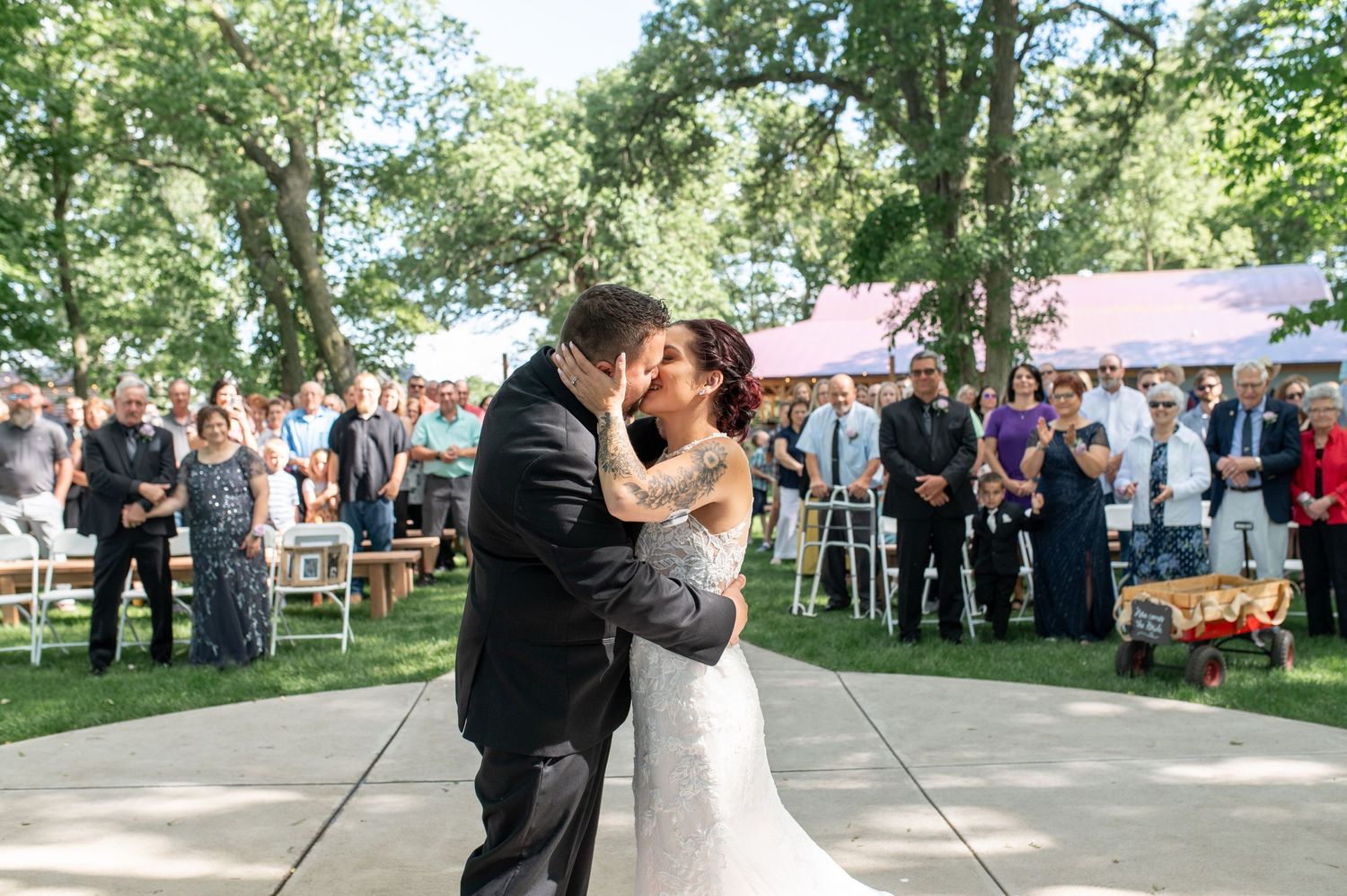 Wedding couple dancing outdoors on a paved area surrounded by trees and guests.