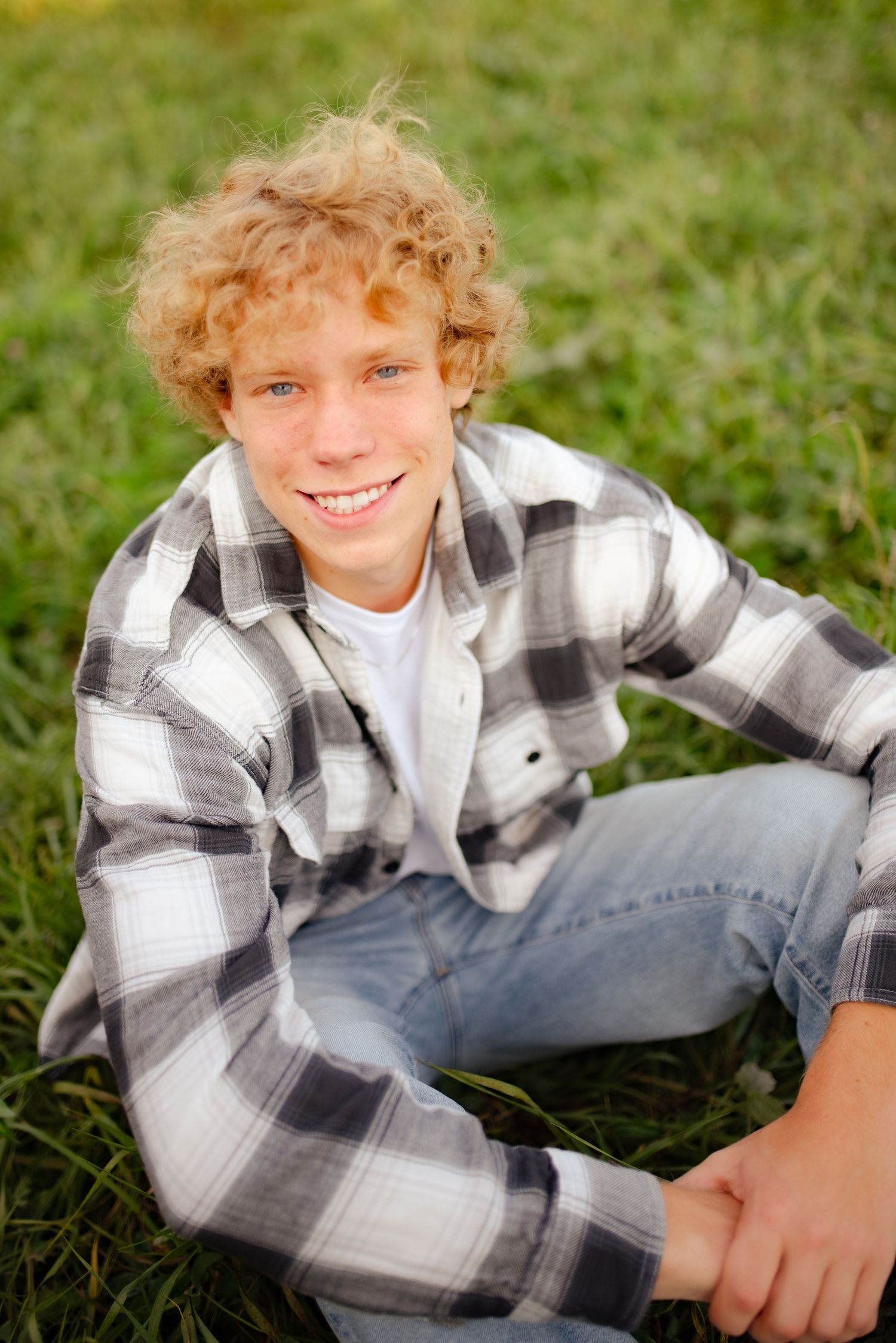 Young person in black and white plaid flannel shirt sitting casually on grass outdoors.