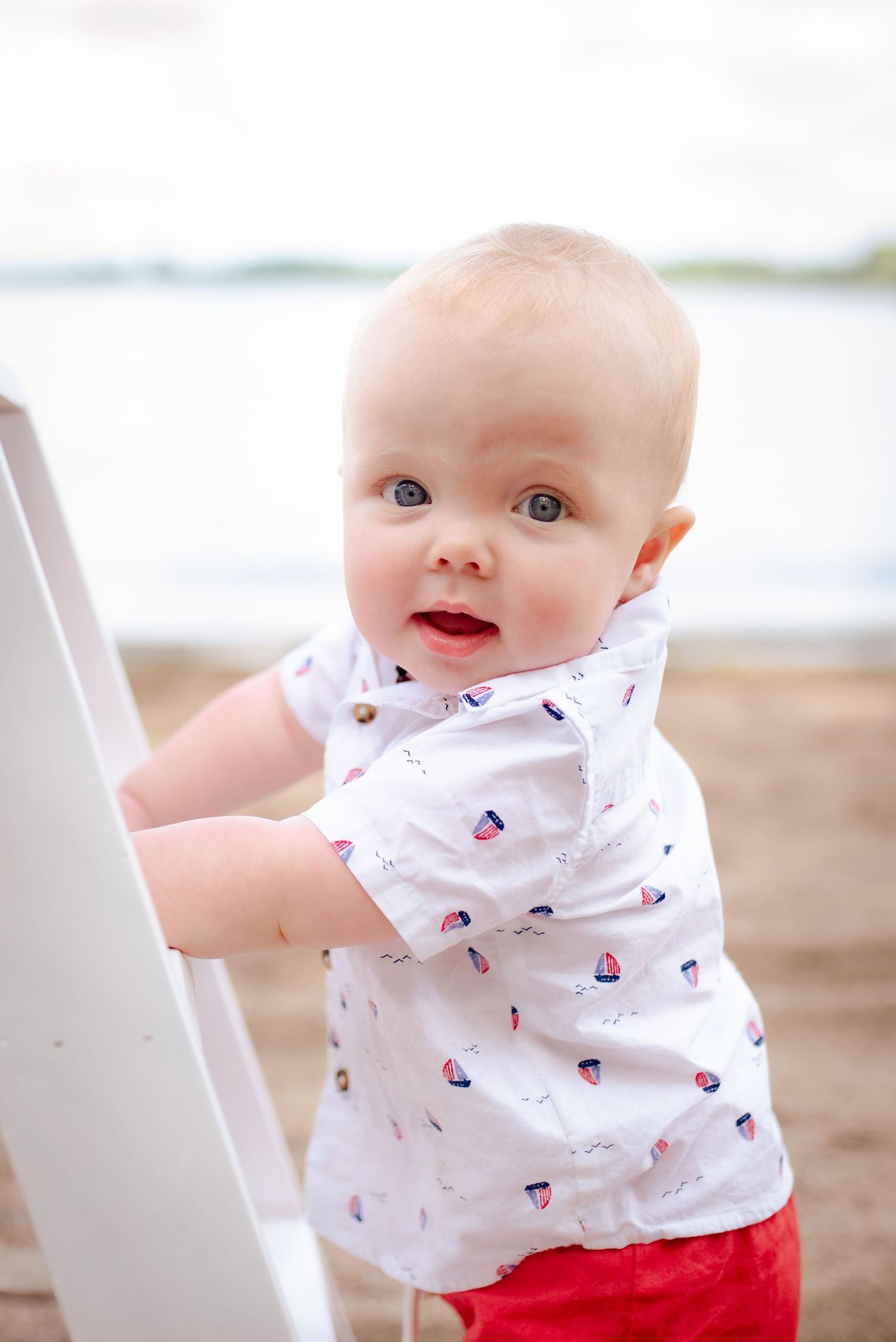 A baby in a white patterned shirt and red shorts stands at the beach holding onto a chair.