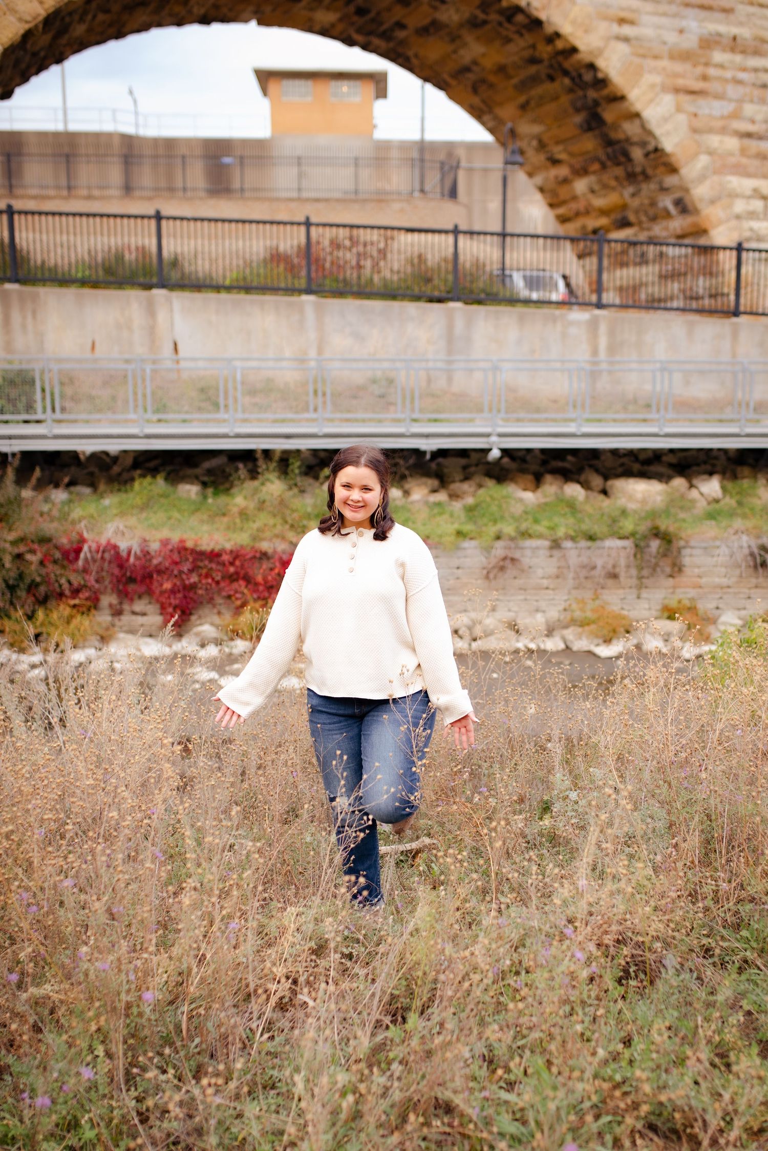A person in white sweater walks through tall grass beneath a stone arch bridge.