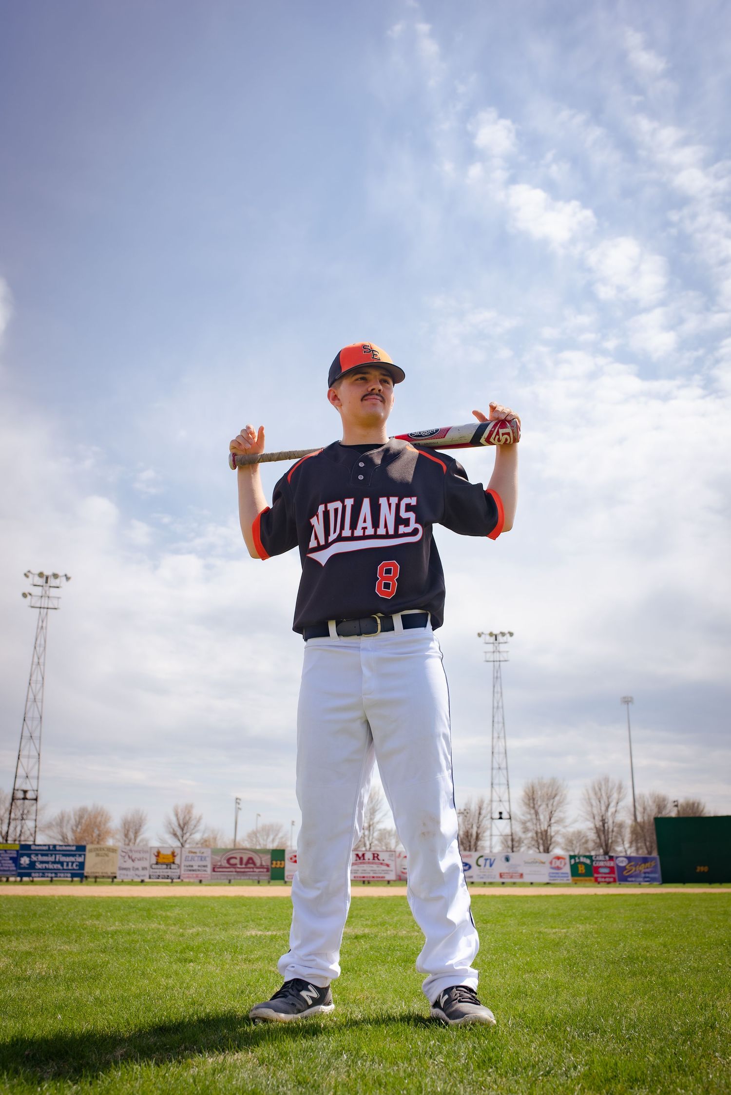 Baseball player in Indians uniform poses with bat on shoulders against cloudy sky.