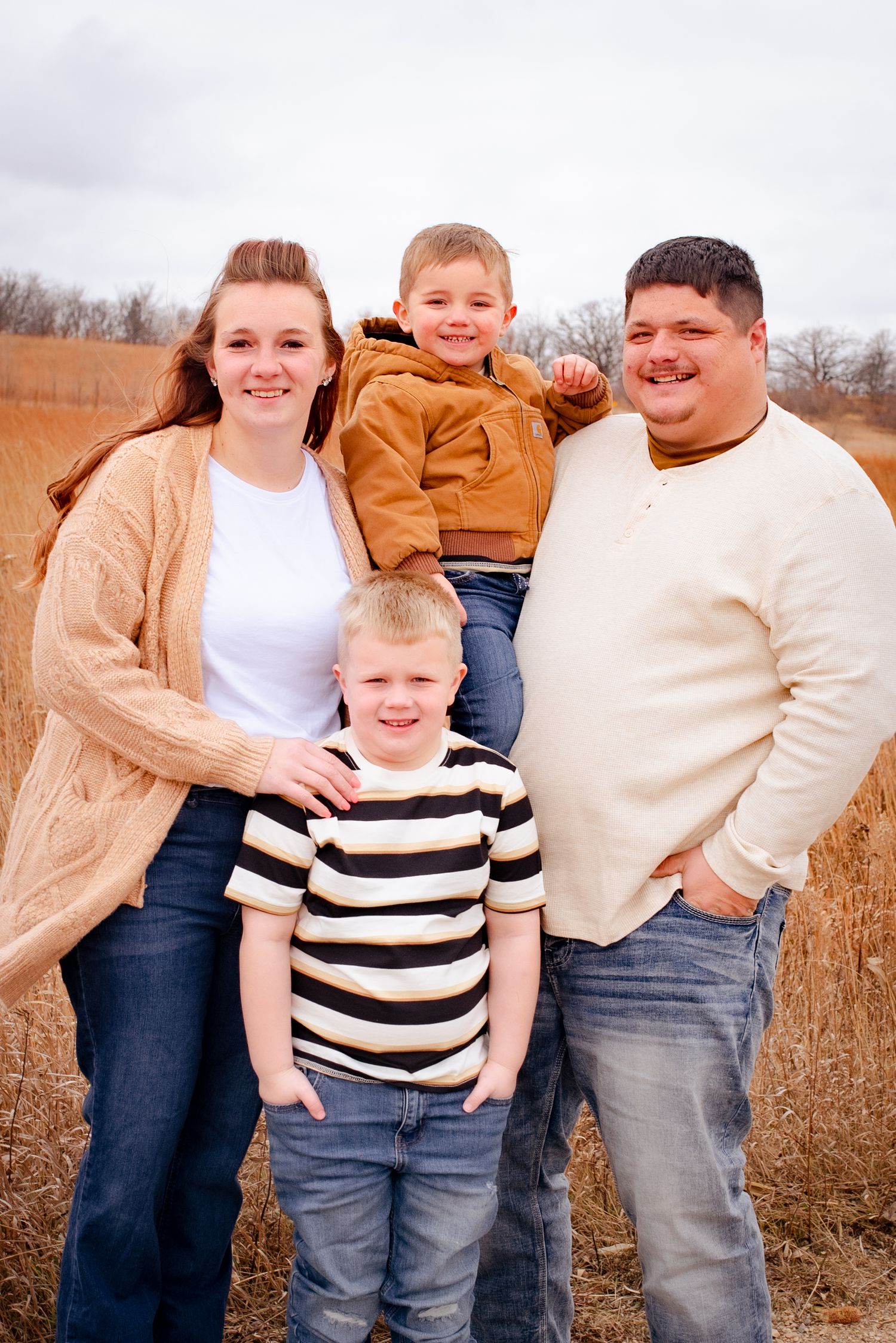 Family of four poses together in casual attire against autumn field backdrop.