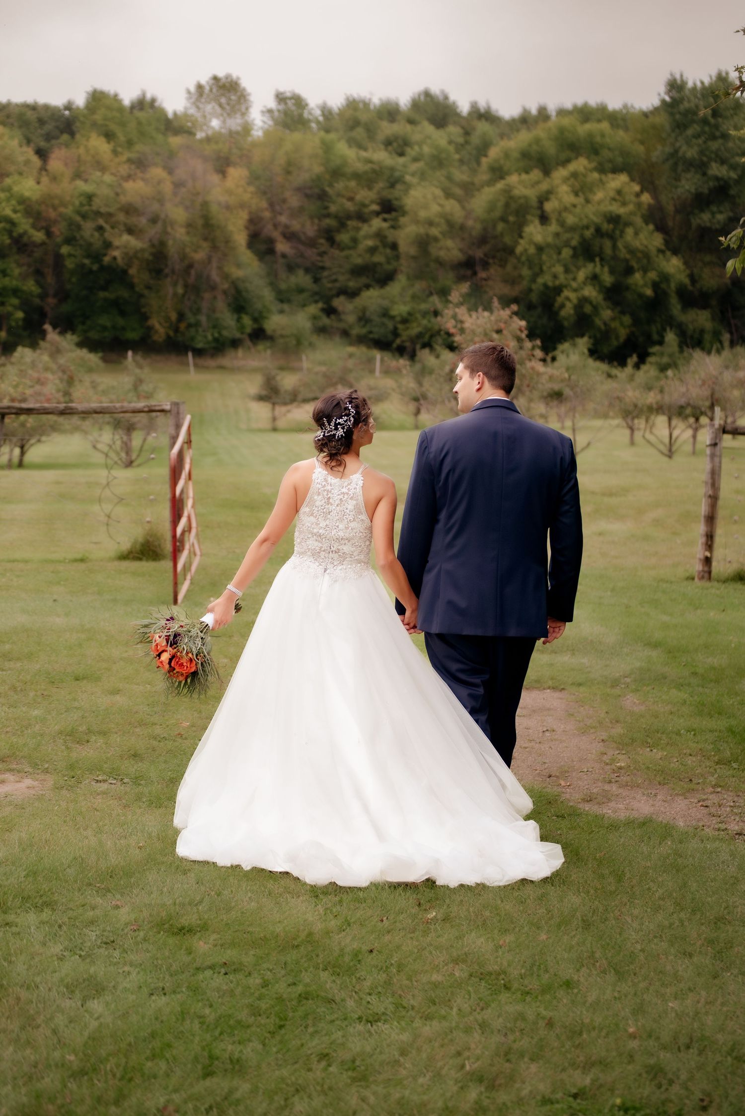 Bride in flowing white gown and groom in navy suit walking together through a scenic orchard.