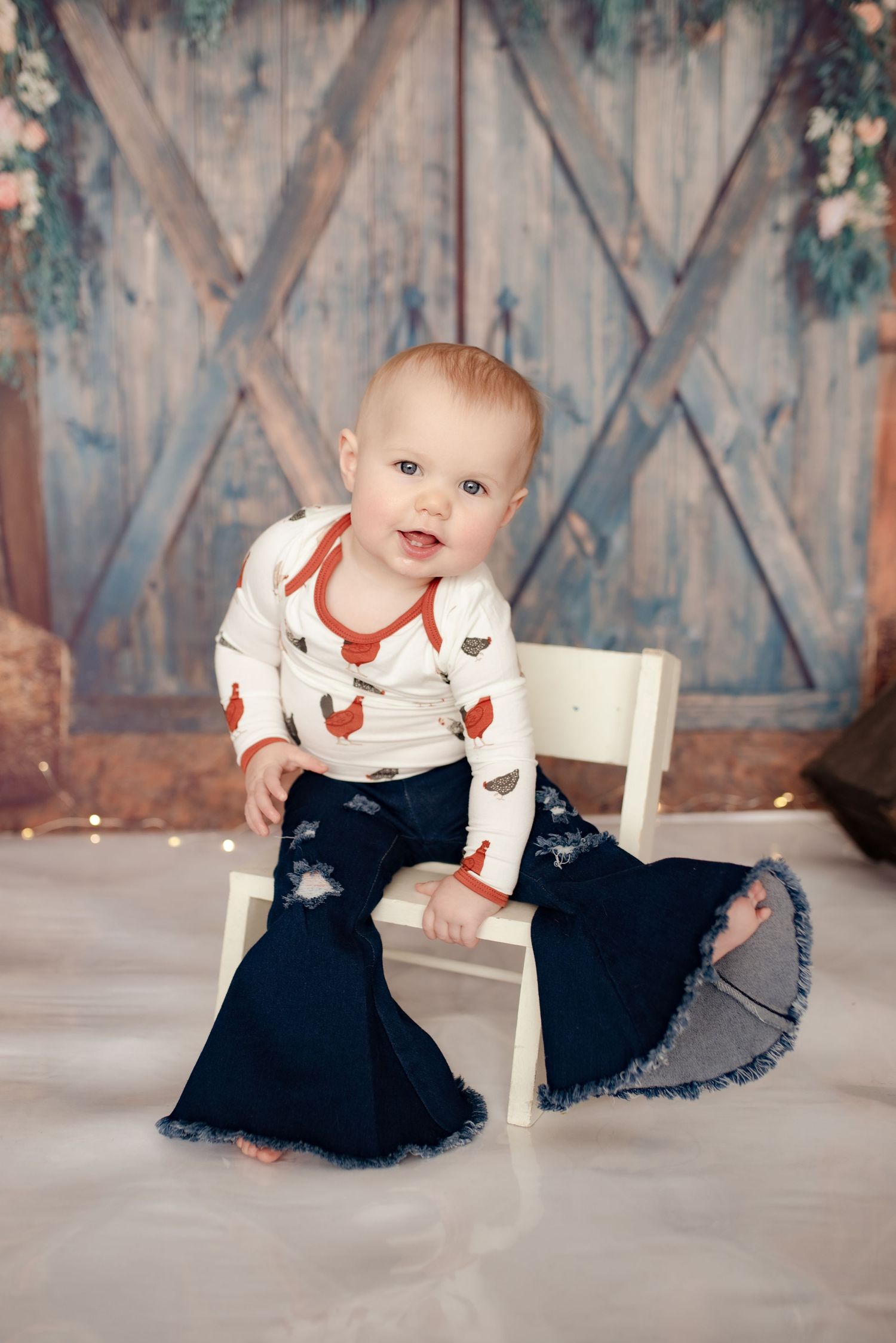 Baby wearing heart print shirt sits on white chair against rustic blue barn door background.