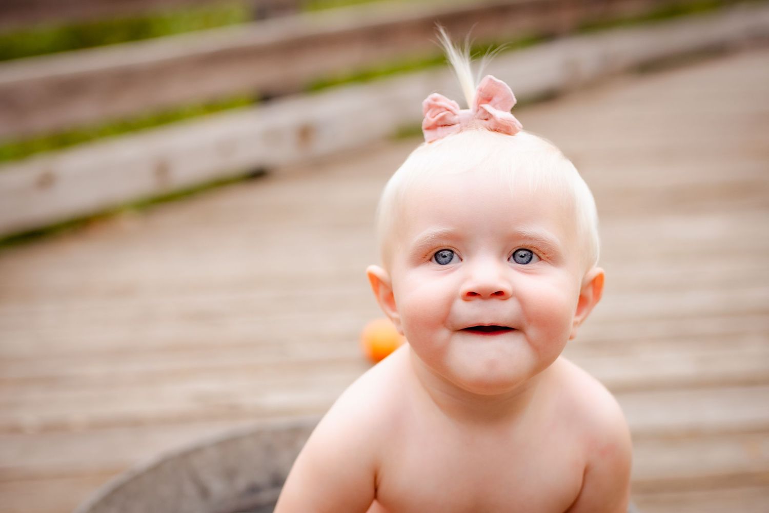 Baby with pink bow smiles while sitting on wooden boardwalk during outdoor photoshoot.