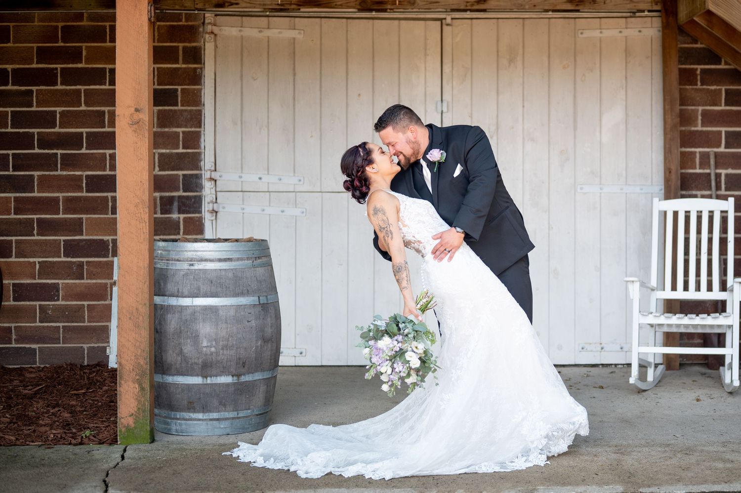 A wedding couple shares an intimate kiss in front of a rustic barn door with a vintage barrel nearby.
