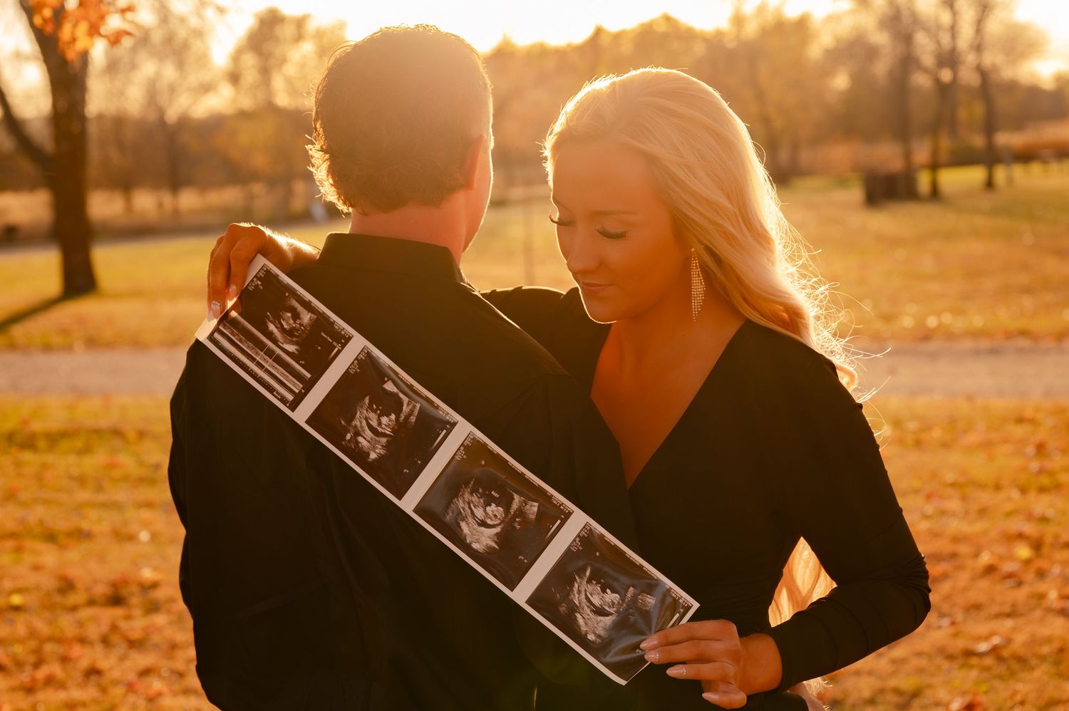 A couple holds up ultrasound photos during a sunset photoshoot in a park with autumn leaves on the ground.