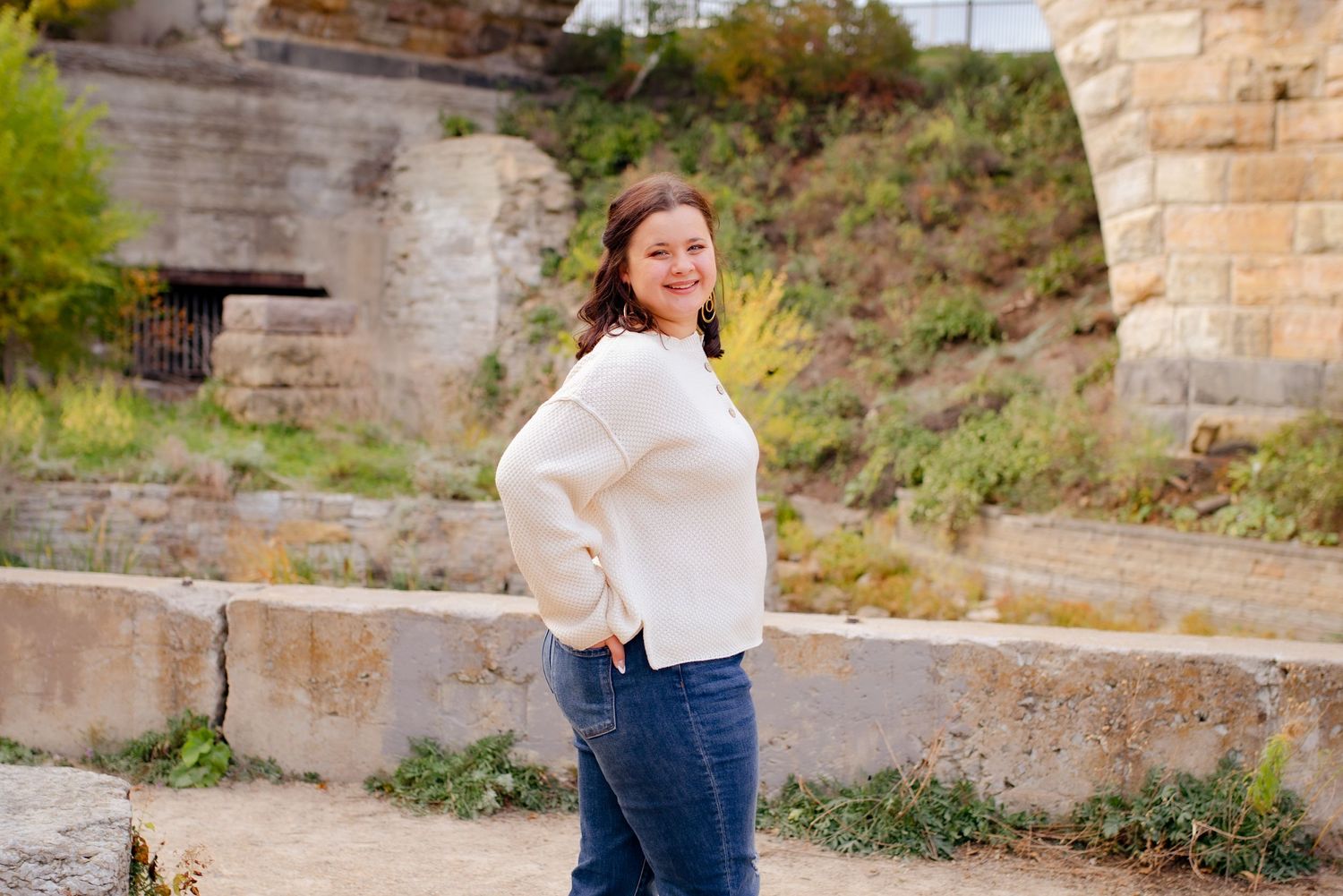 Person in white sweater poses near stone steps with bridge architecture in background.