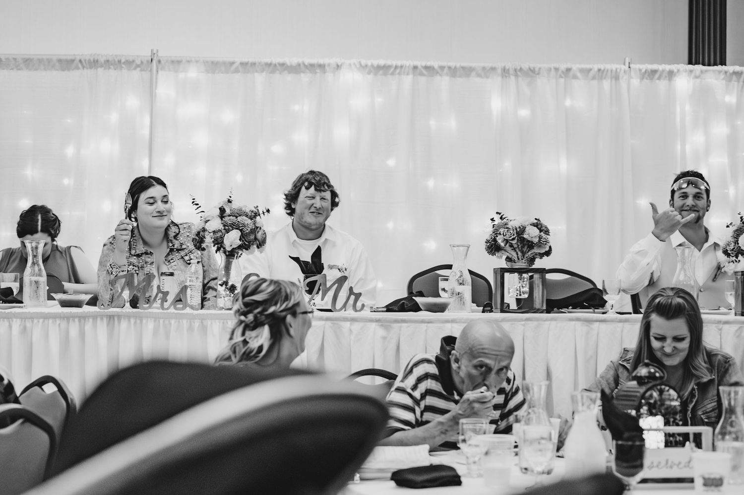 Black and white photo of wedding reception guests seated at tables with decorative lighting in background.