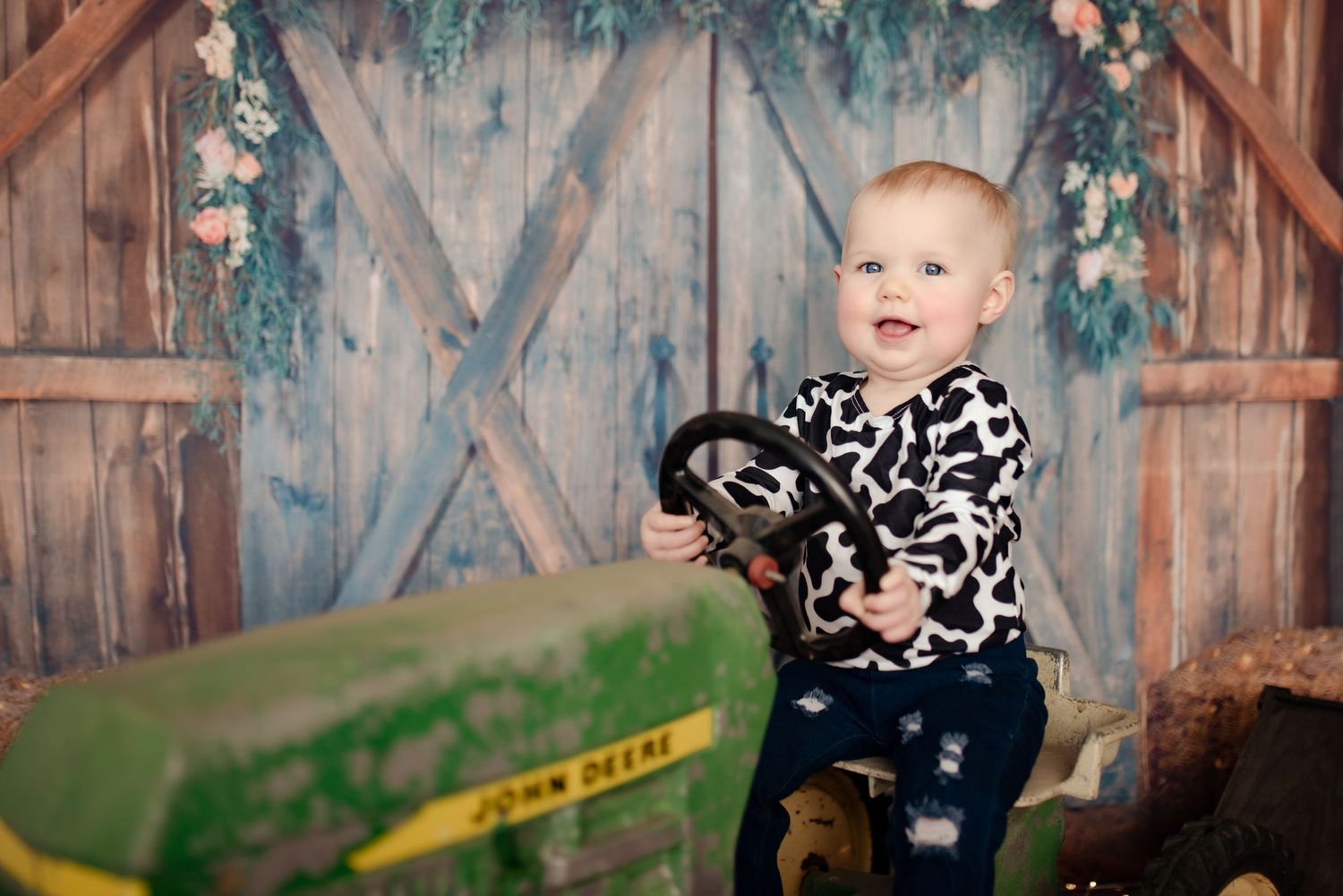 Baby wearing cow print sweater plays with toy tractor in rustic barn setting.